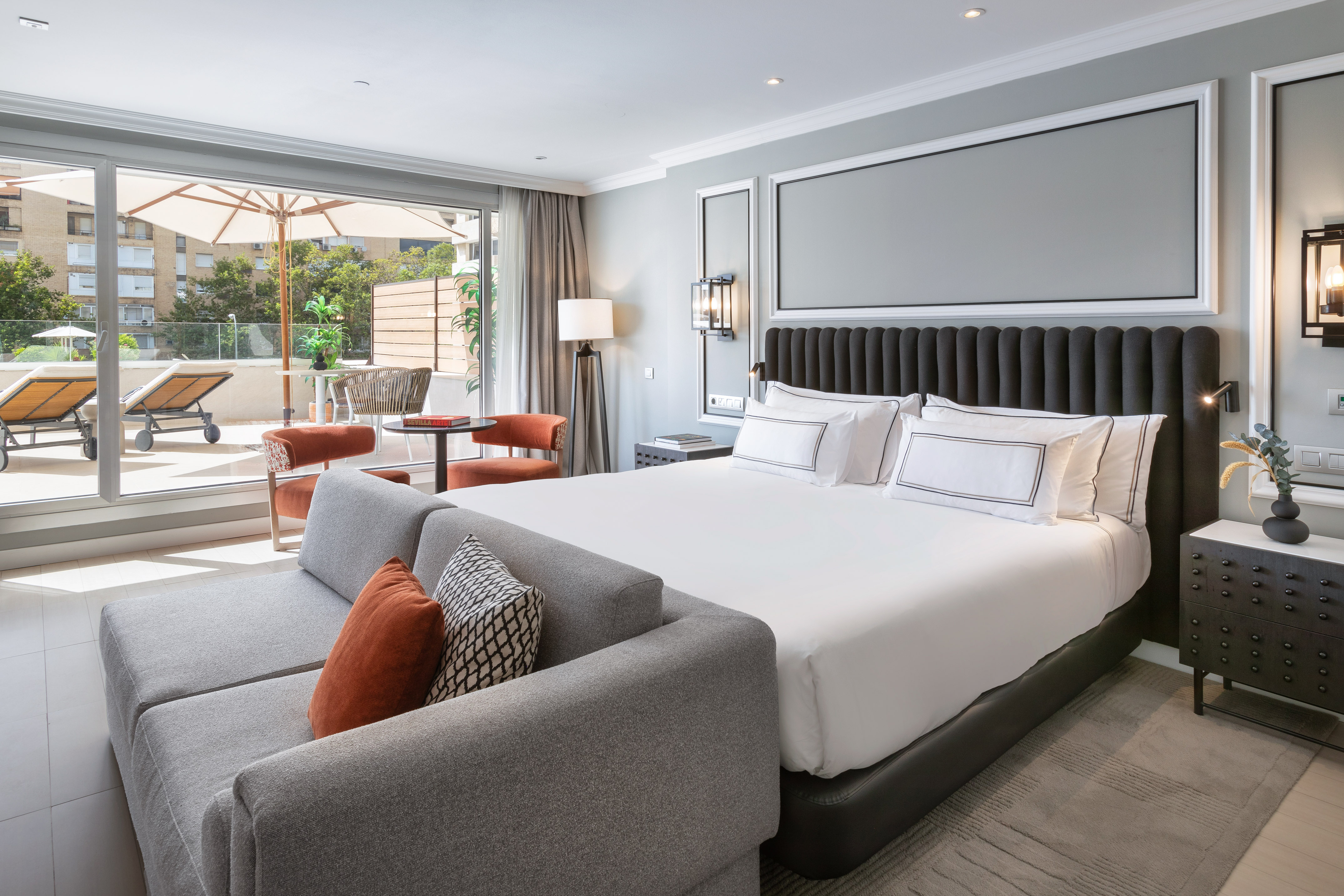 Modern hotel bedroom with king bed, grey sofa, and private terrace.