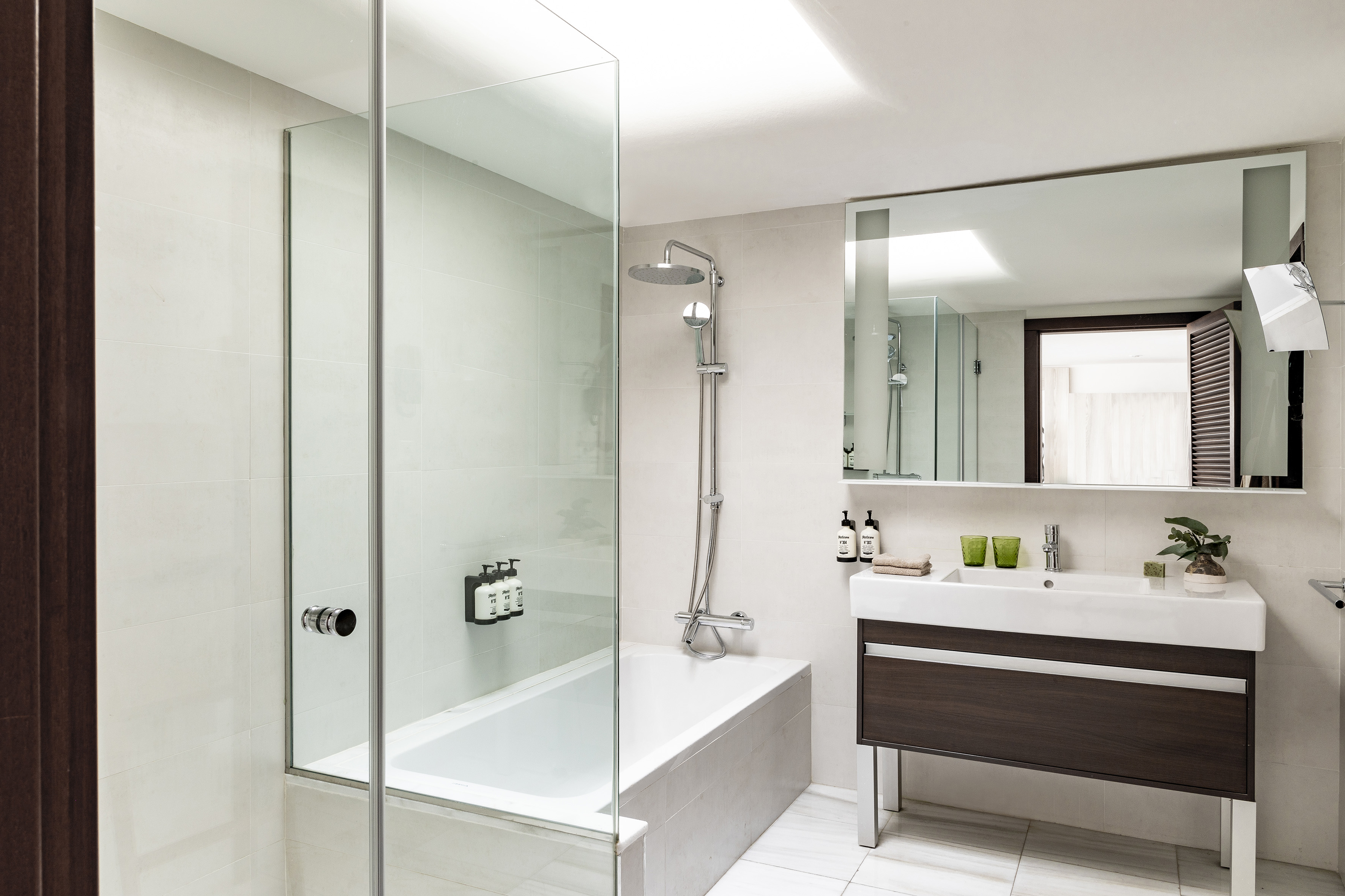 Bright modern hotel bathroom with glass shower, bathtub, and wood vanity.
