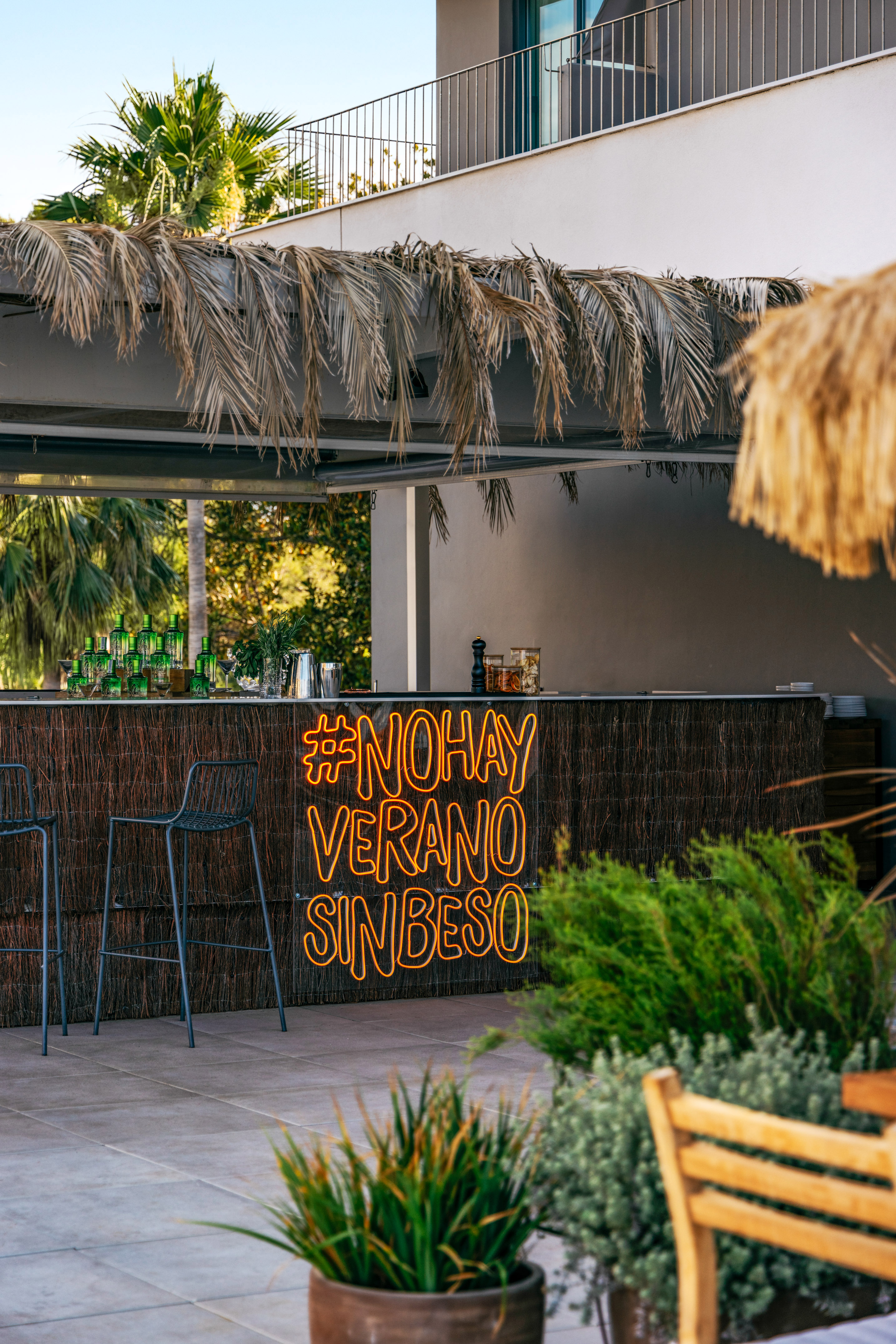 Tropical resort bar with "#NOHAY VERANO SINBESO" neon sign, palm fronds.