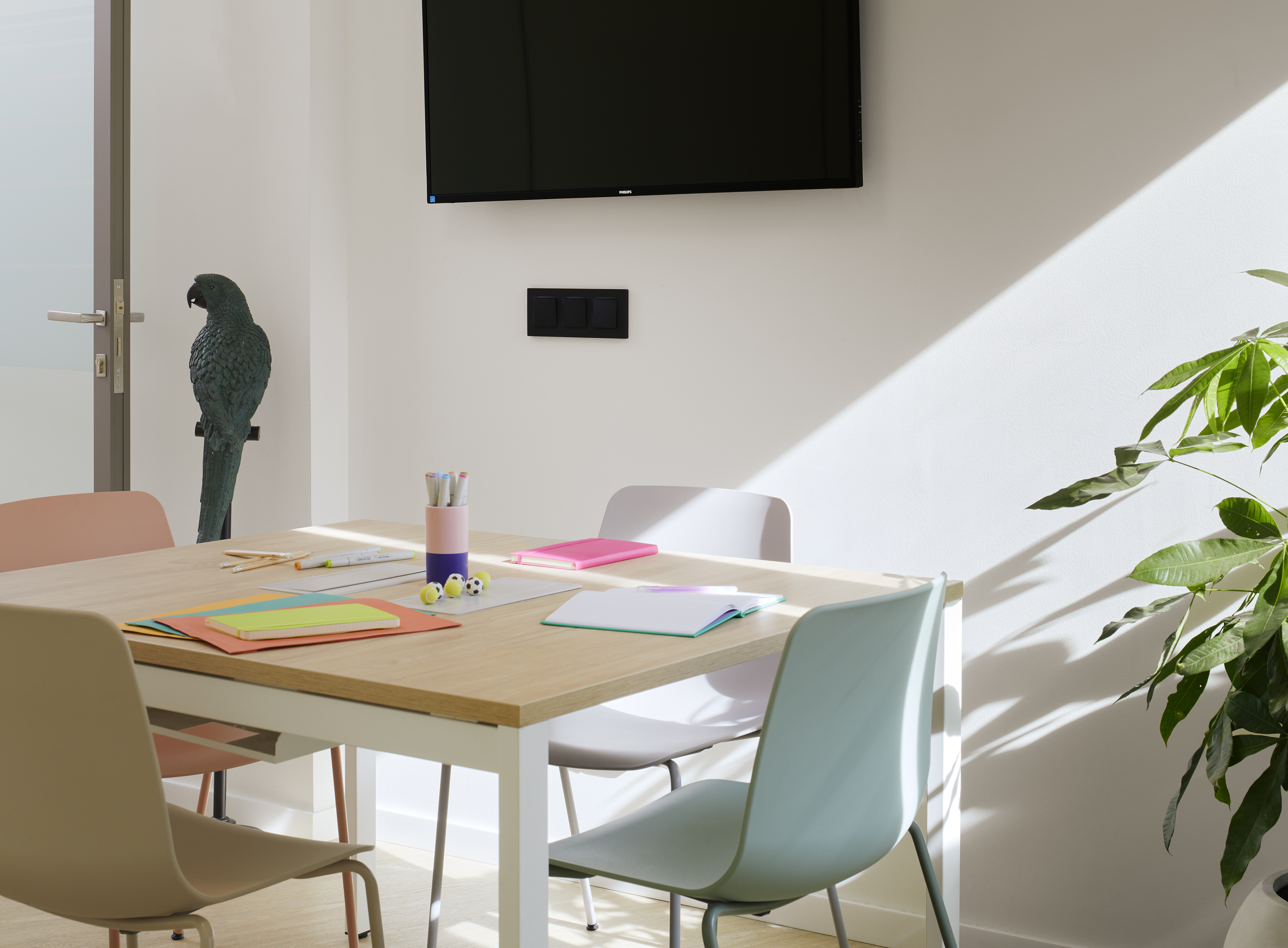 Modern meeting room with colorful chairs, creative supplies on wood table, TV, and plant.