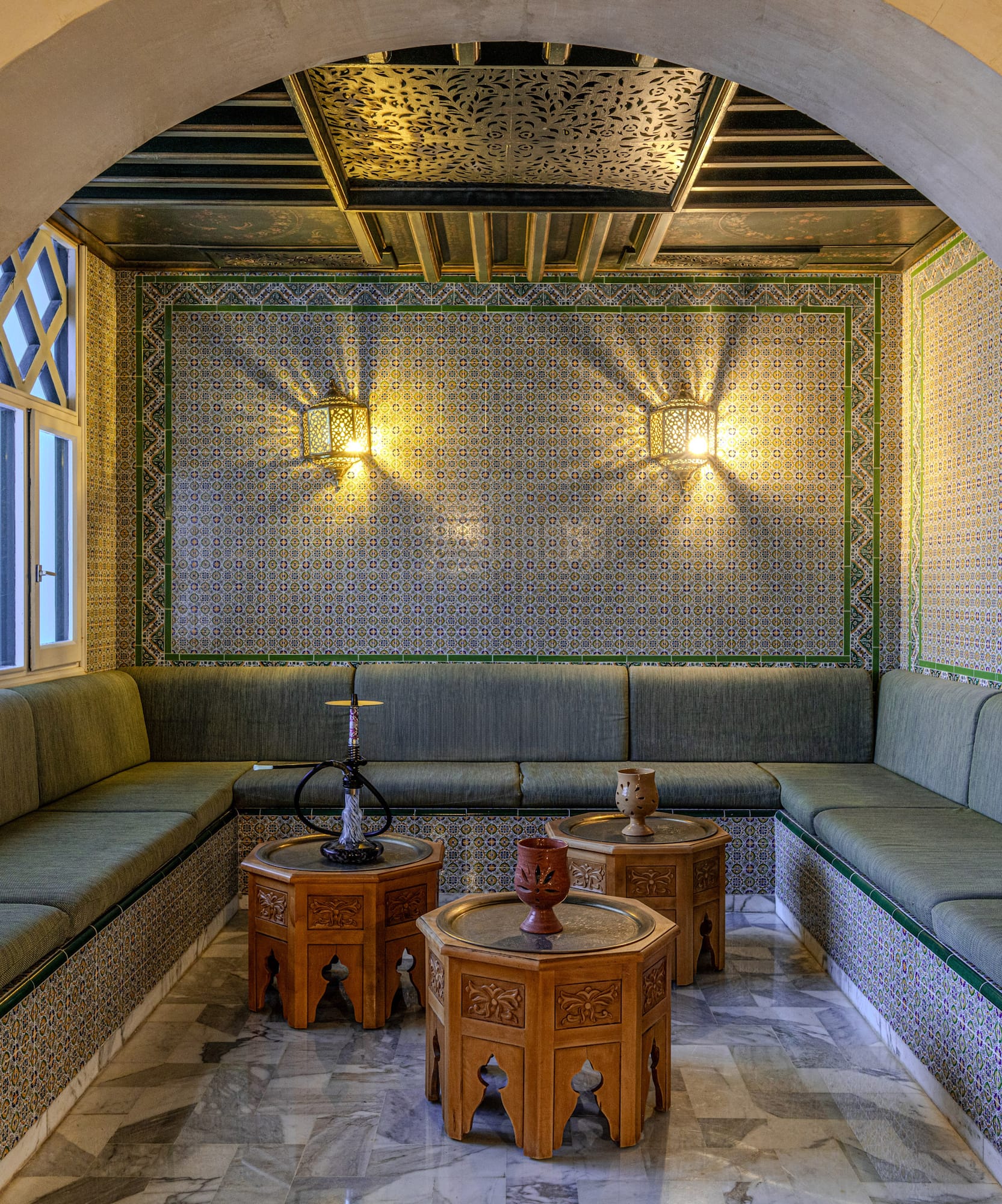 Salón árabe tradicional con sofás verdes, azulejos coloridos y mesas de madera tallada.