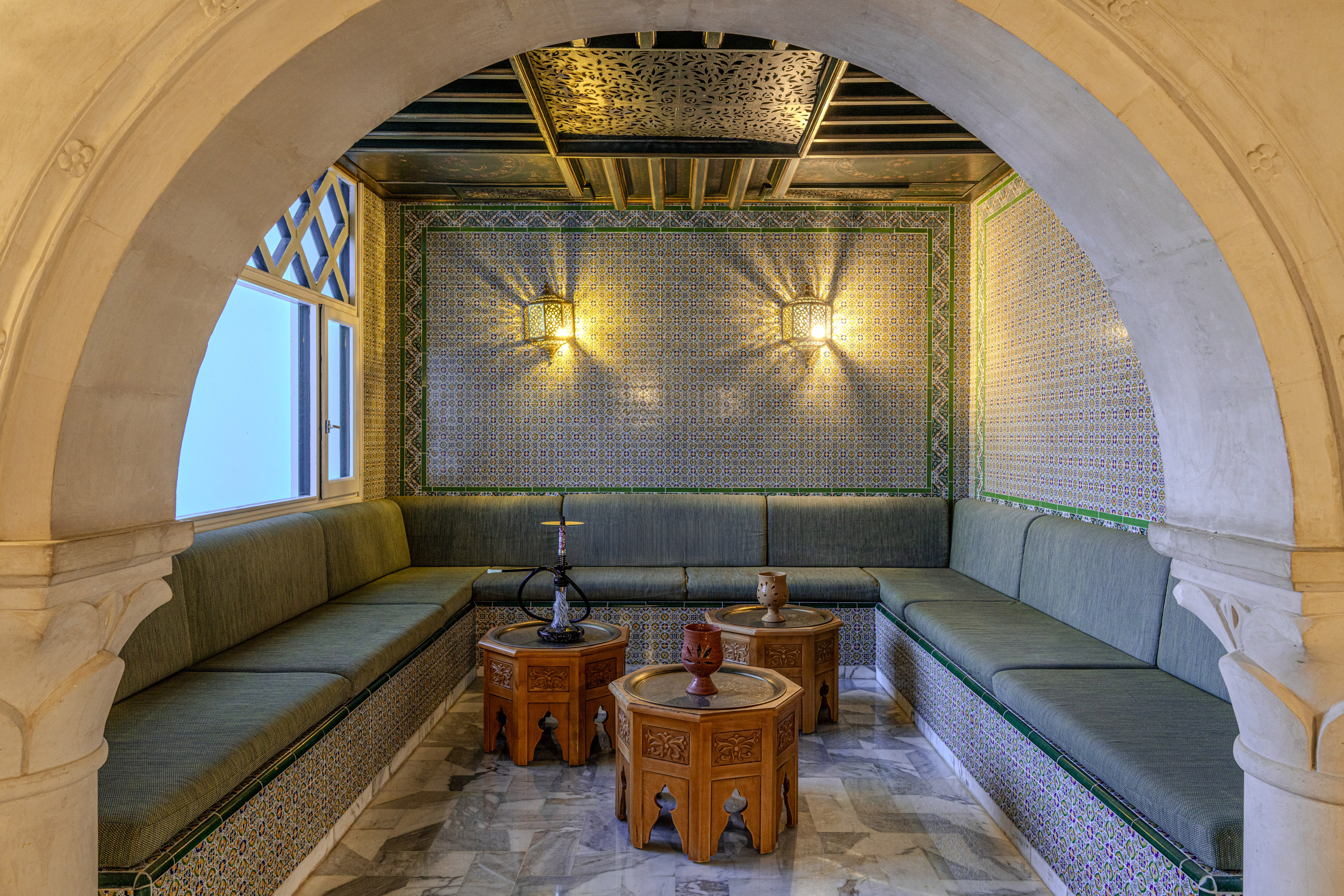 Salón árabe tradicional con sofás verdes, azulejos coloridos y mesas de madera tallada.