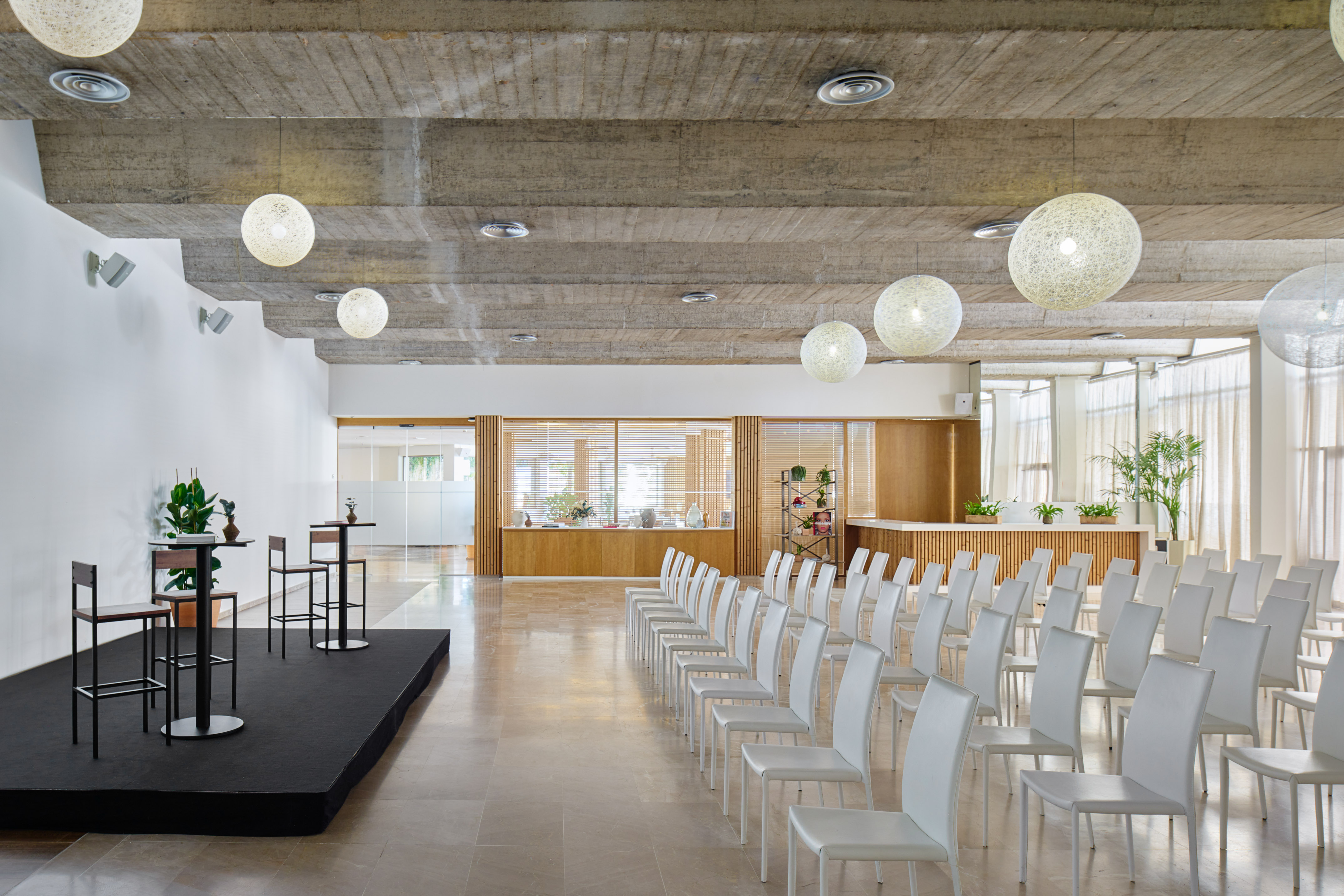 Modern event hall with white chairs, stage, concrete ceiling, and globe lighting.