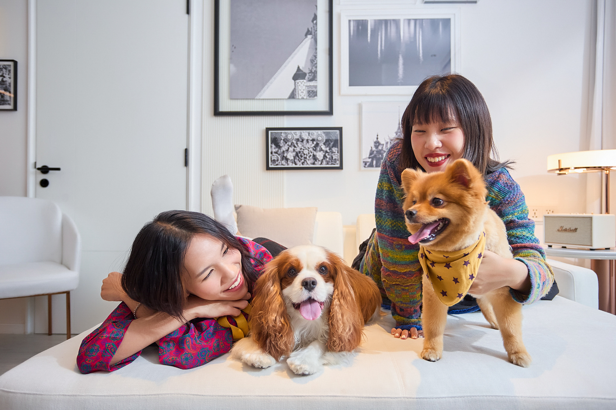 Smiling women with Cavalier and Pomeranian dogs on a white sofa.