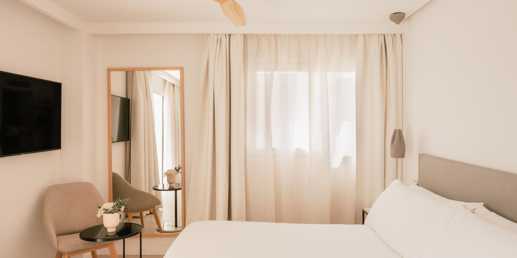 Modern minimalist hotel bedroom with white bed, wooden fan, TV, and mirror.