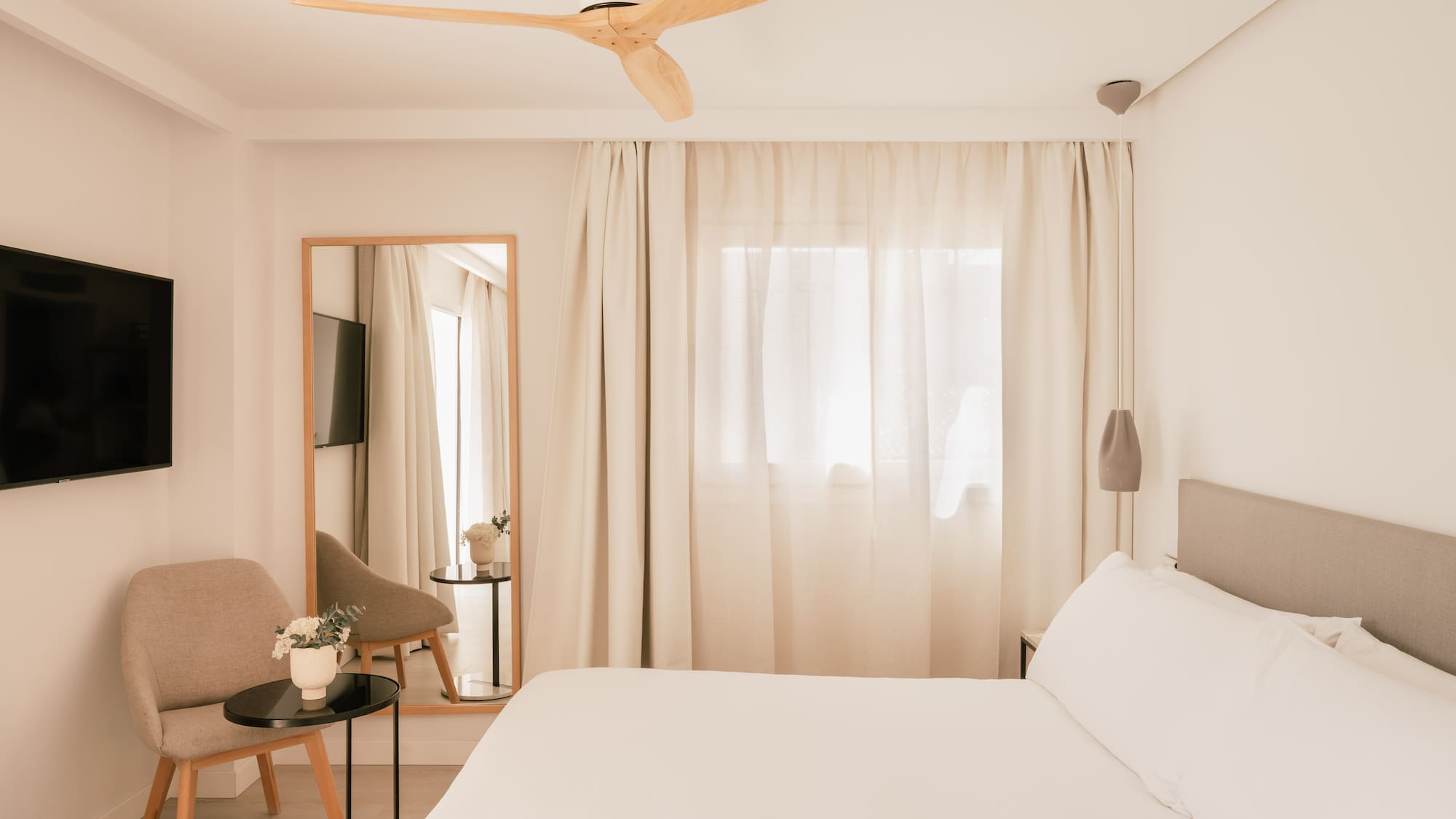 Modern minimalist hotel bedroom with white bed, wooden fan, TV, and mirror.