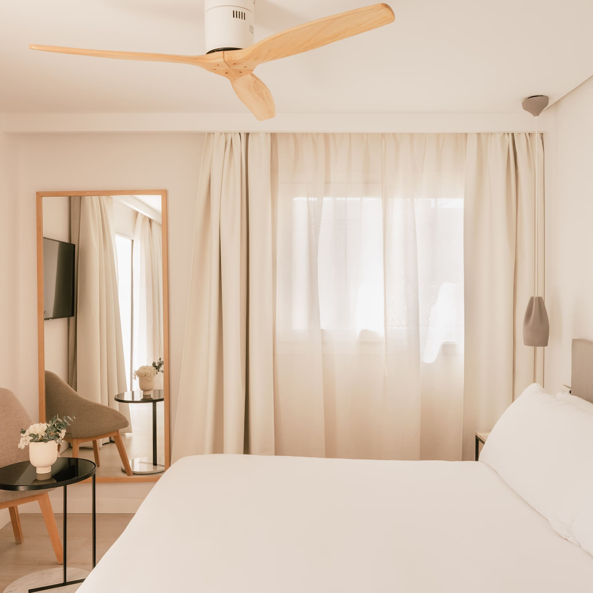 Modern minimalist hotel bedroom with white bed, wooden fan, TV, and mirror.