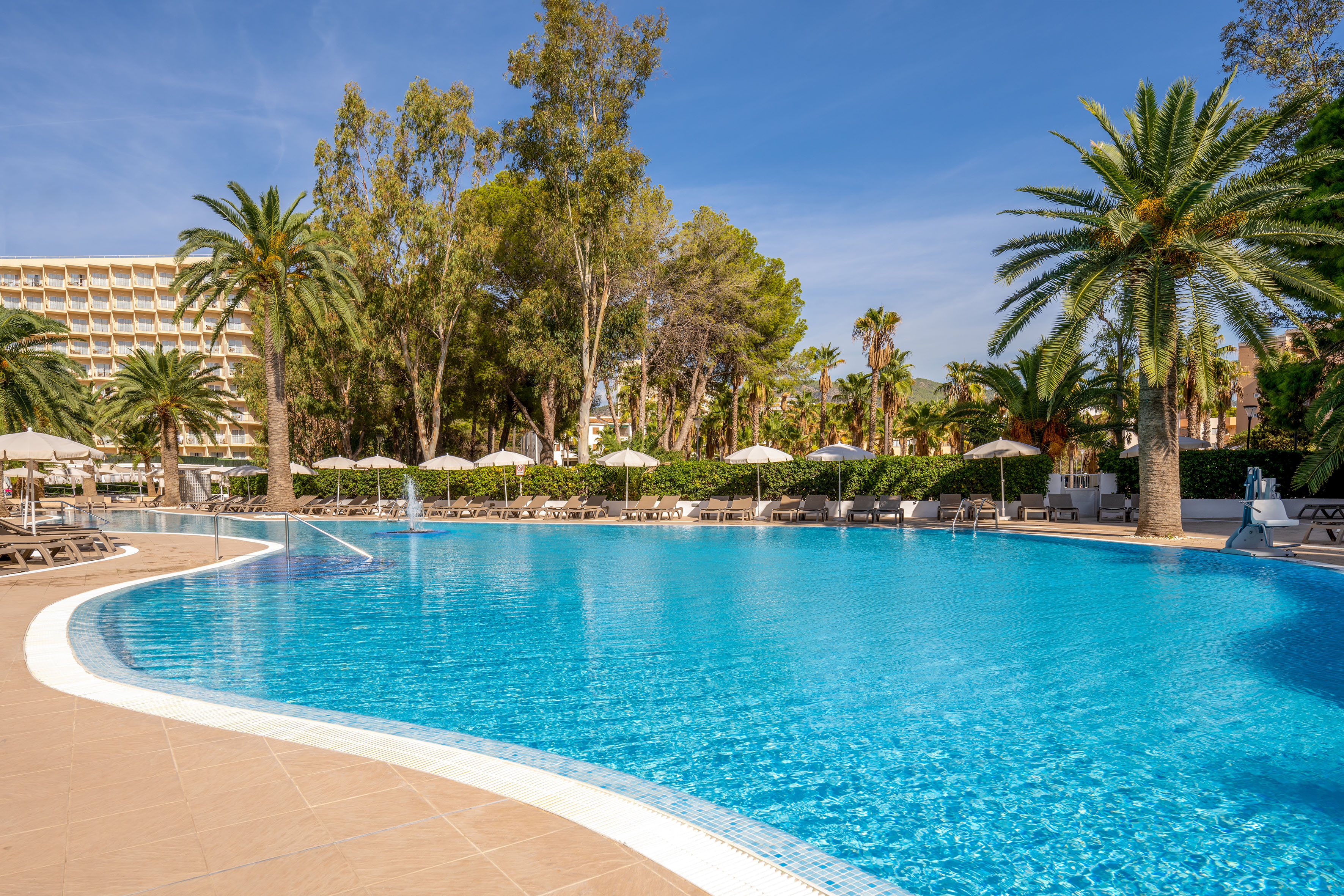 Outdoor resort pool with palm trees, sun loungers, and white parasols.