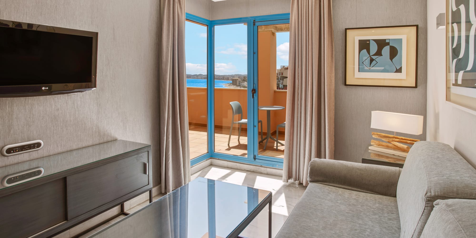 Hotel living room suite with balcony, sea view, gray sofa, and TV.