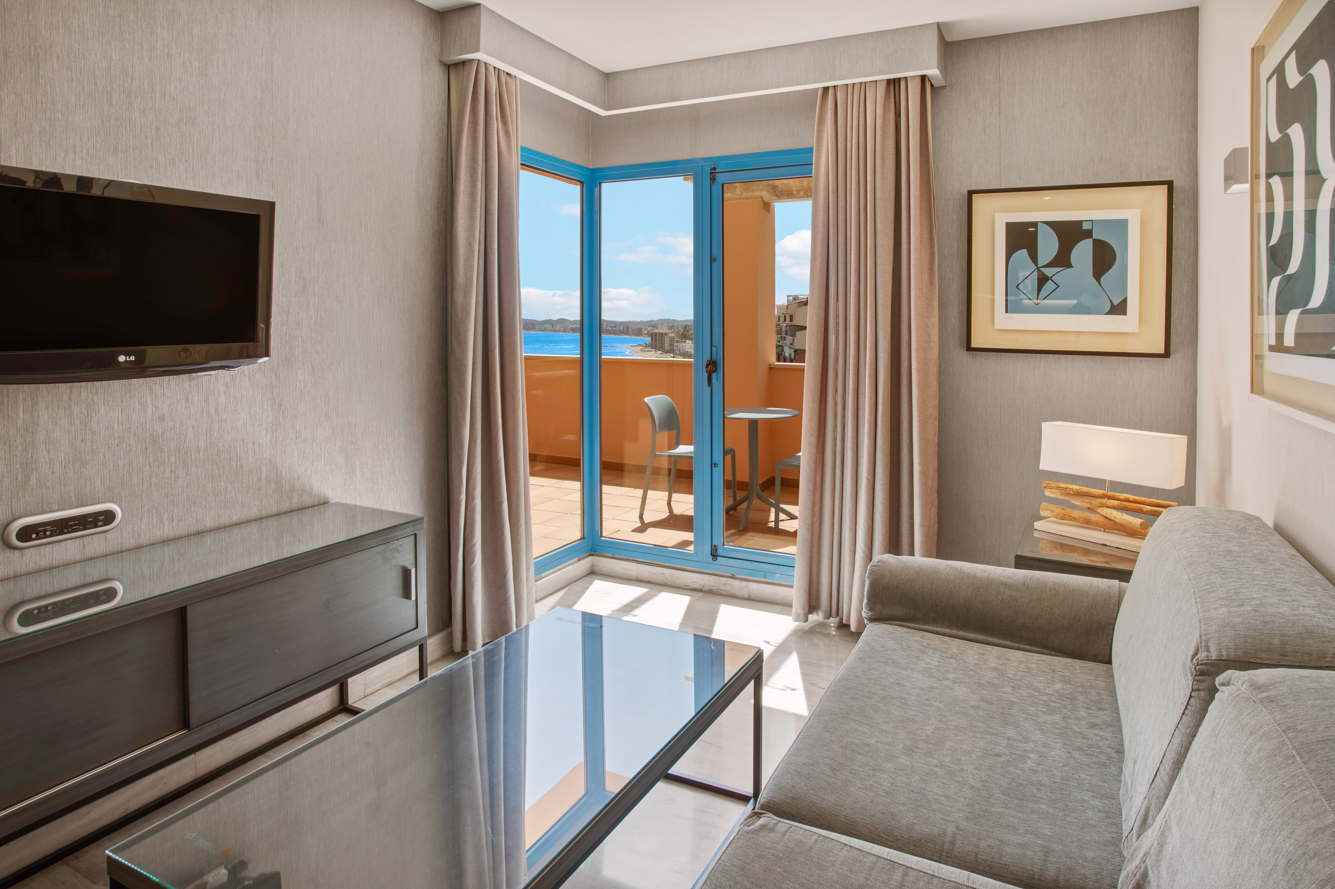 Hotel living room suite with balcony, sea view, gray sofa, and TV.