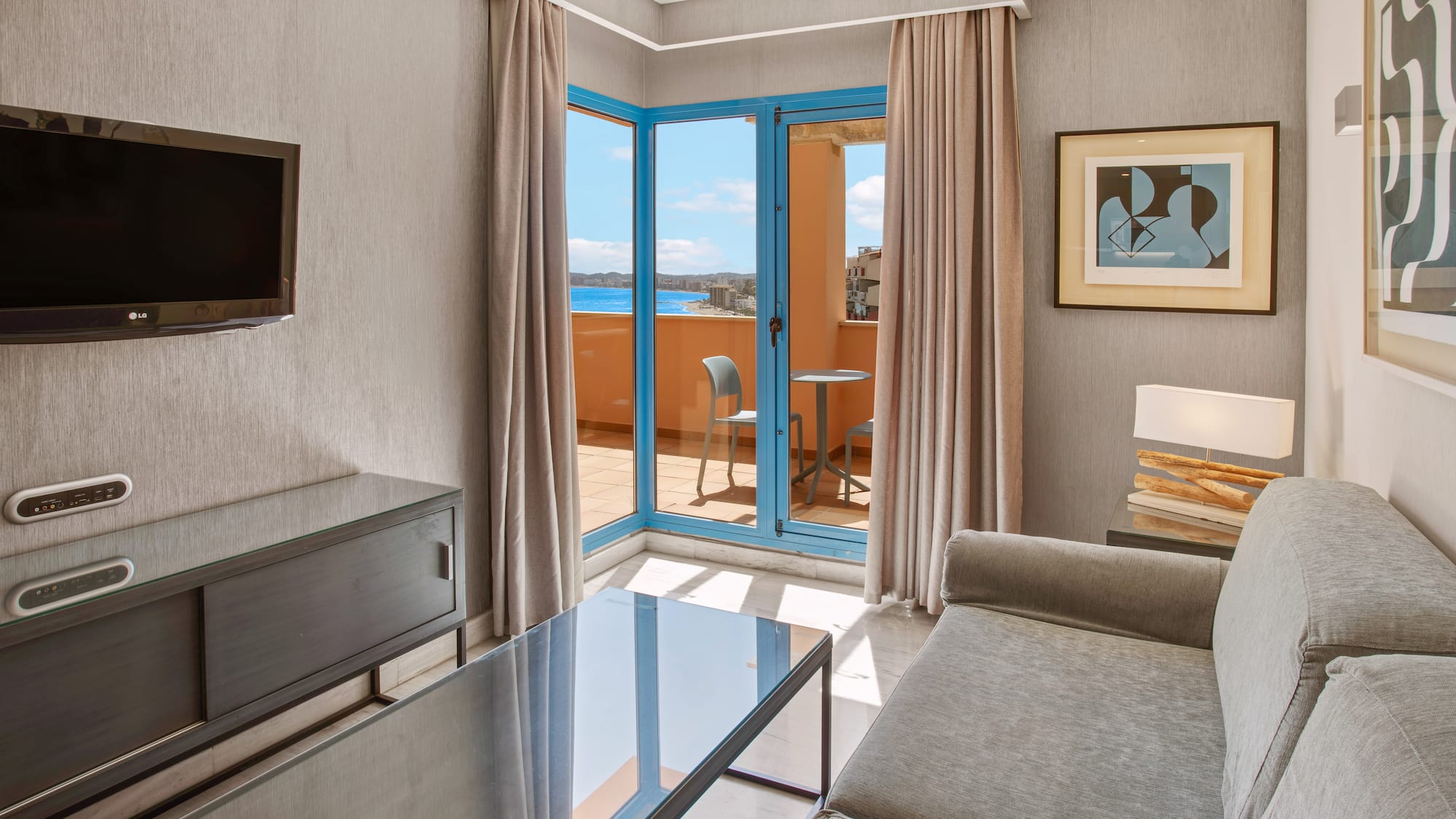 Hotel living room suite with balcony, sea view, gray sofa, and TV.
