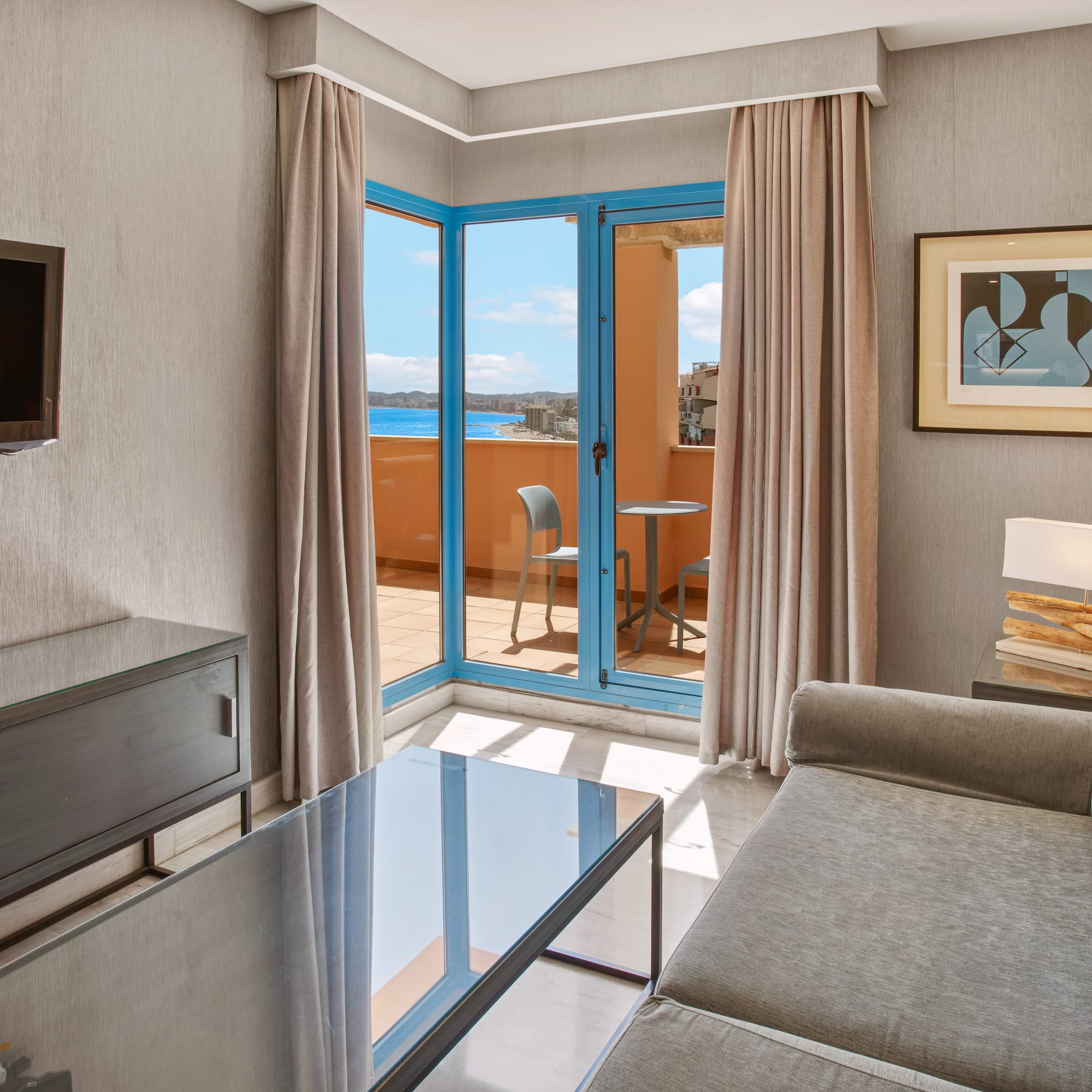 Hotel living room suite with balcony, sea view, gray sofa, and TV.