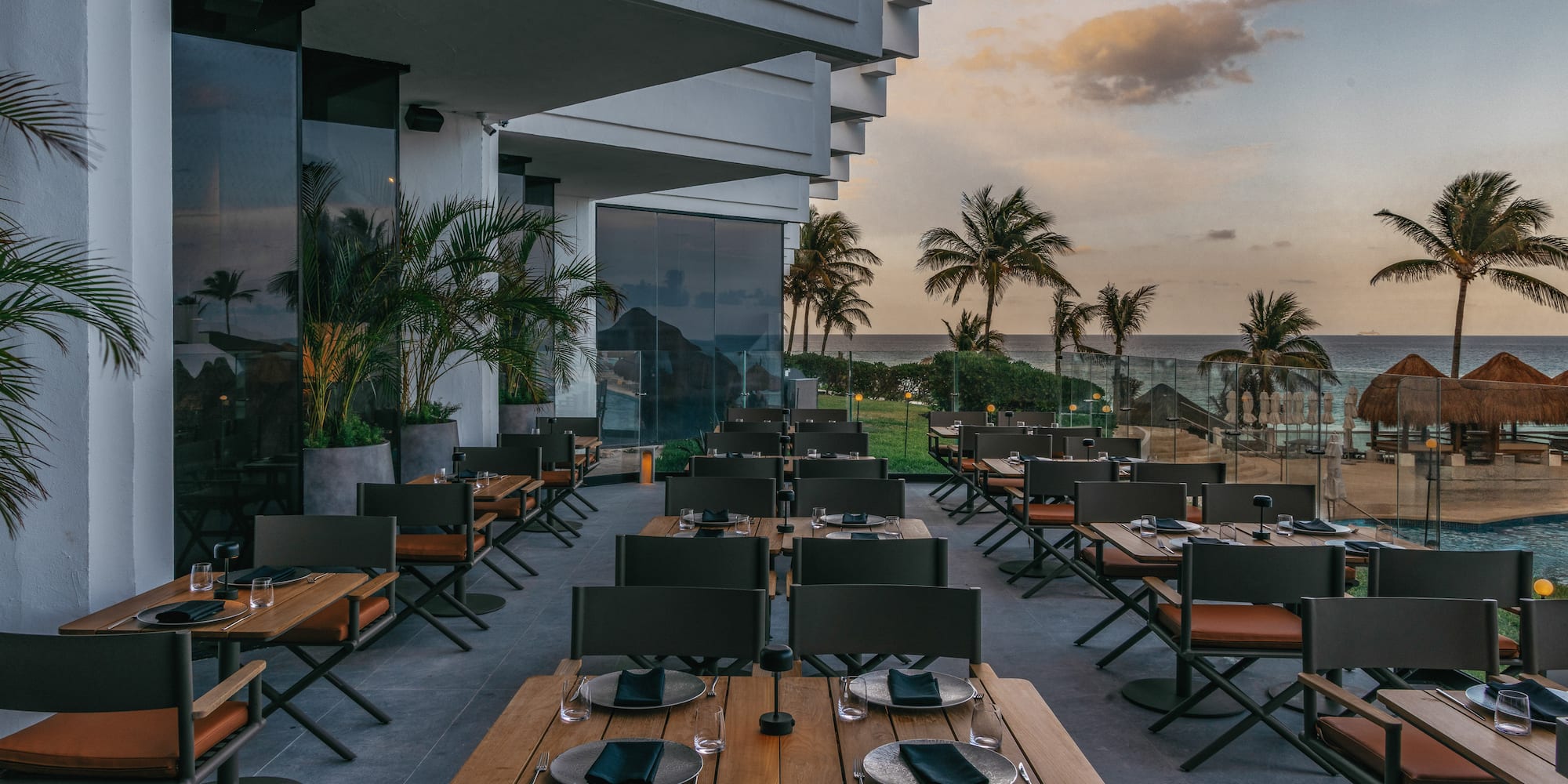 Luxury outdoor restaurant patio with ocean view, palm trees, and sunset.