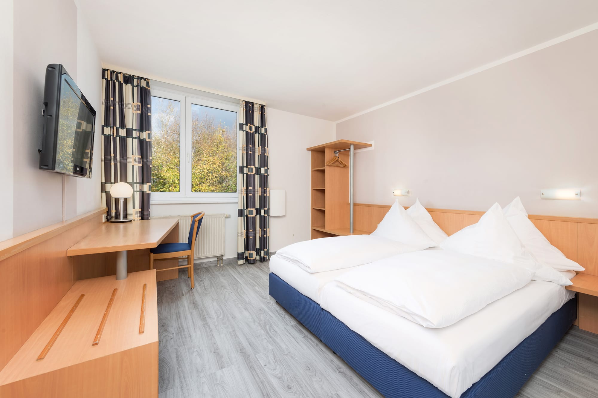 Hotel bedroom with twin beds, desk, TV, and fall foliage outside window.
