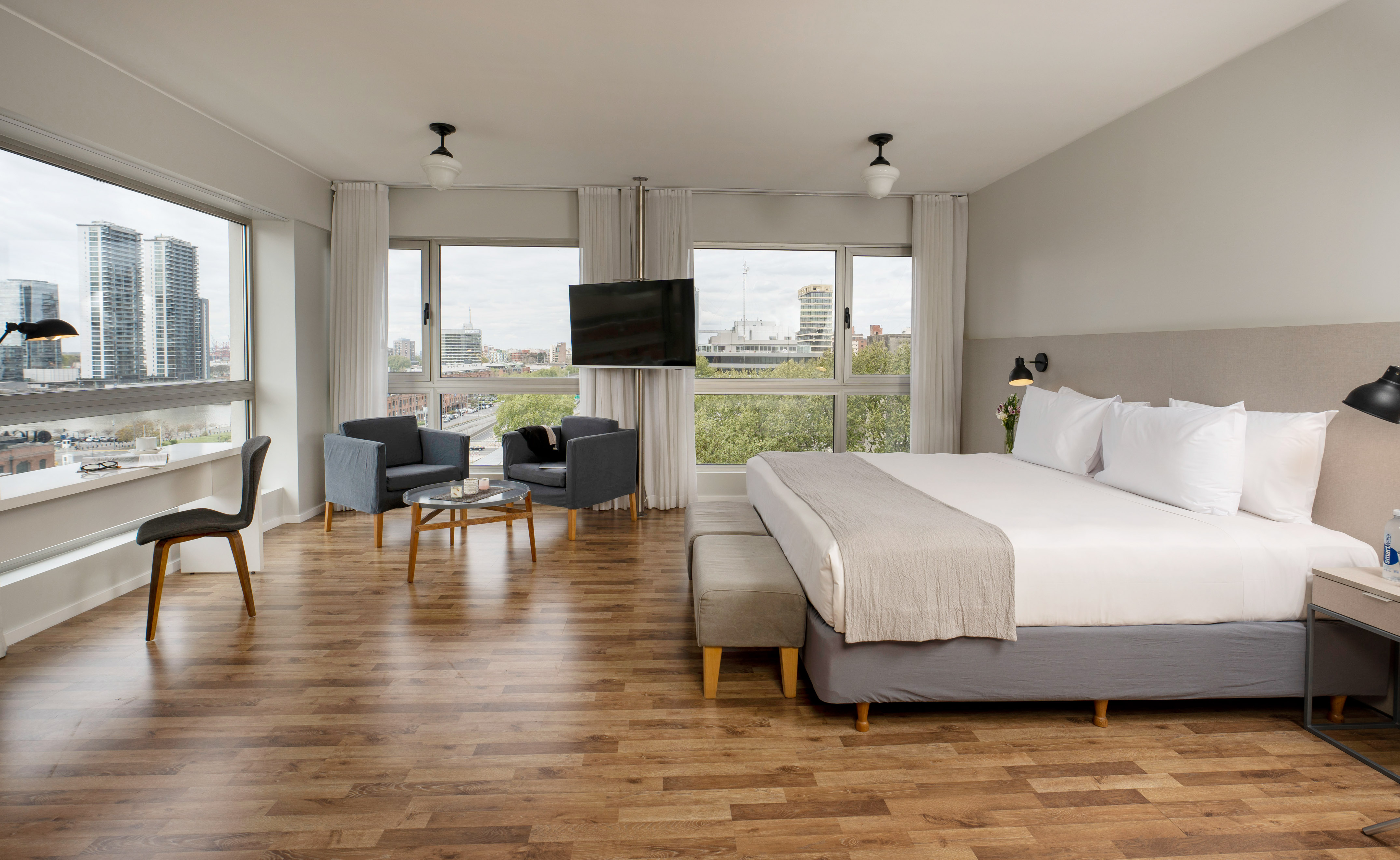 Modern hotel room with city view, king bed, armchairs, and wooden floor.