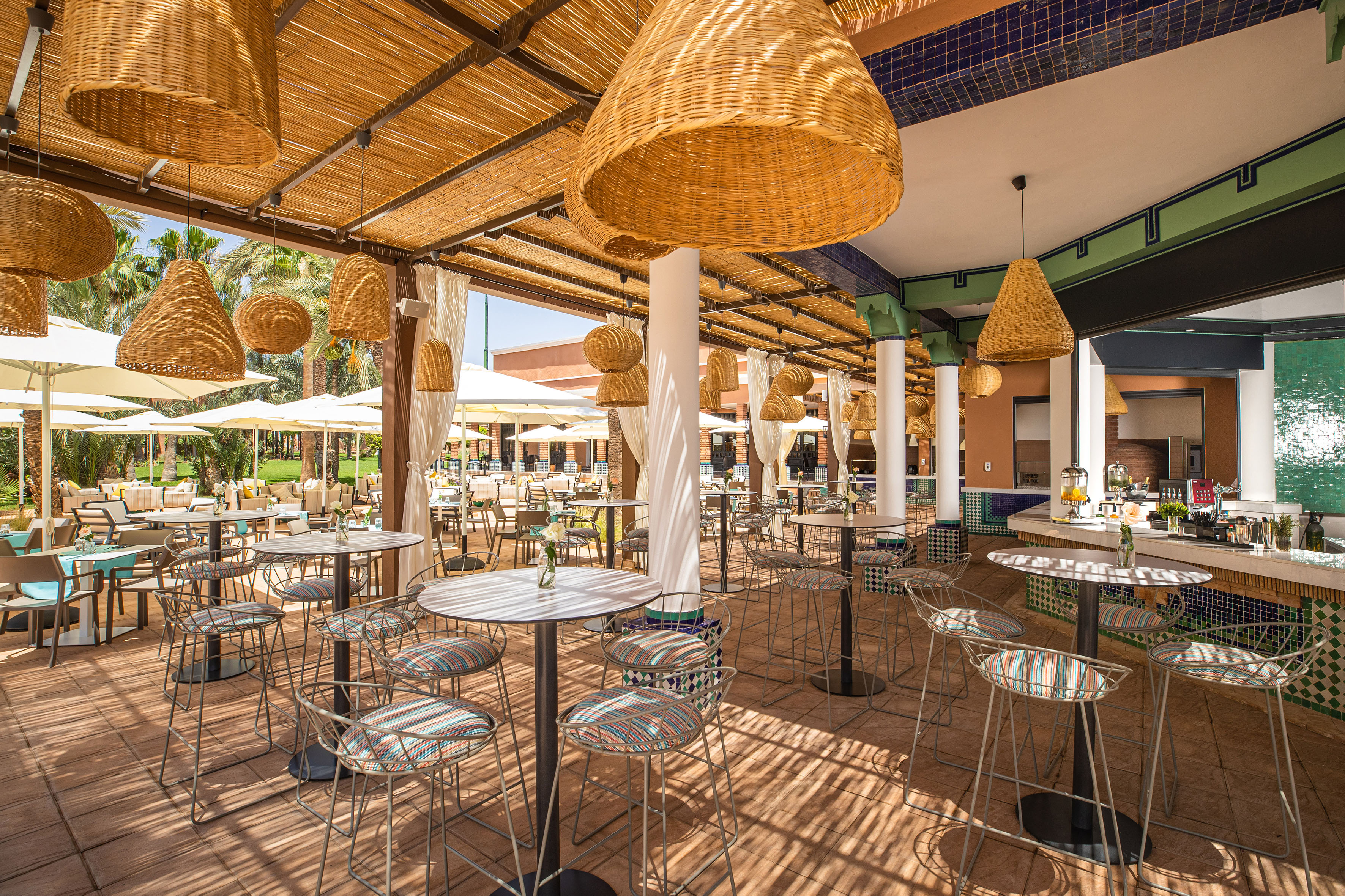 Outdoor resort bar patio with woven straw ceiling, modern stools, and palm trees.