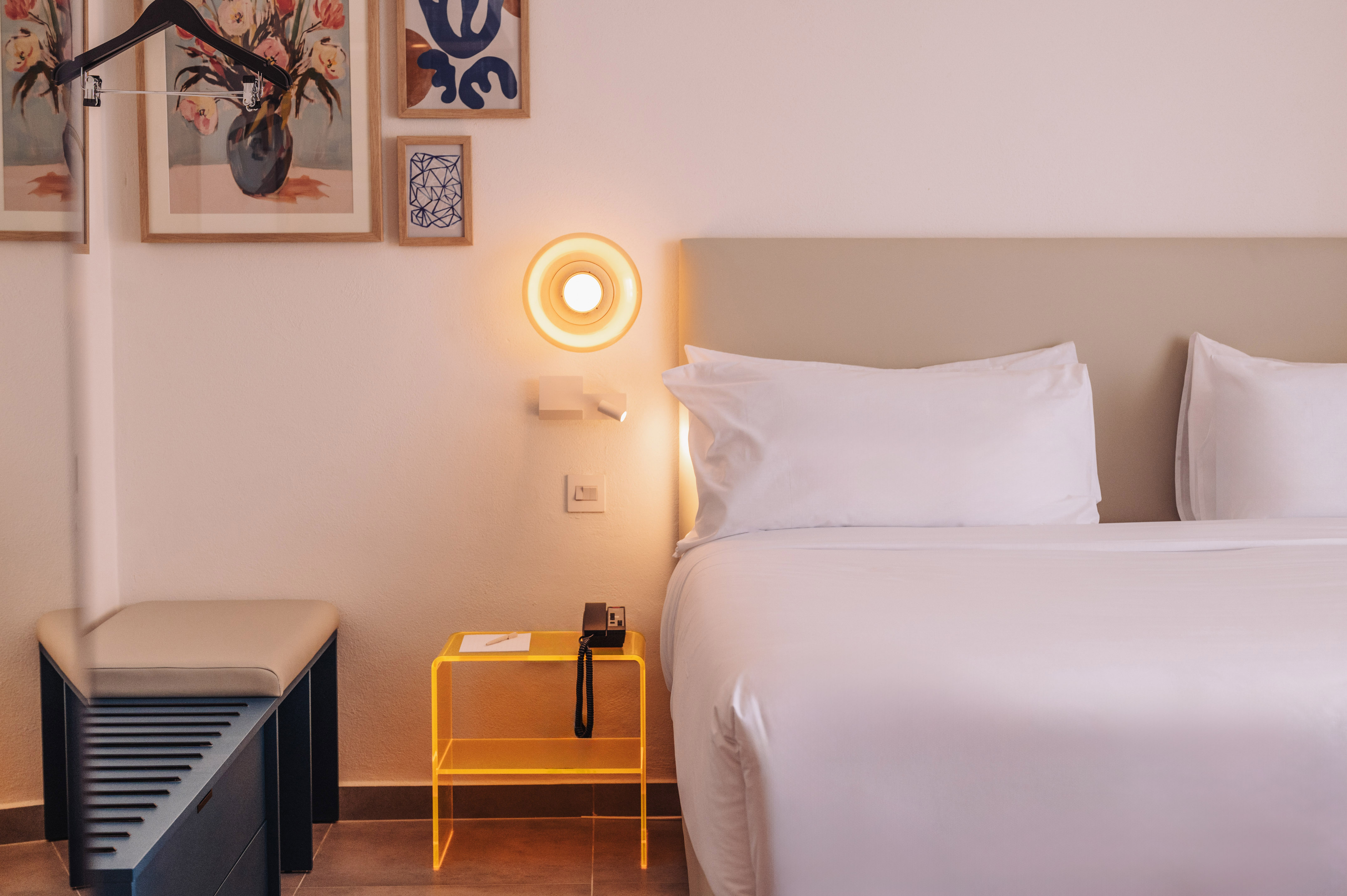 Modern hotel room: white bed, yellow nightstand, wall light, framed art.