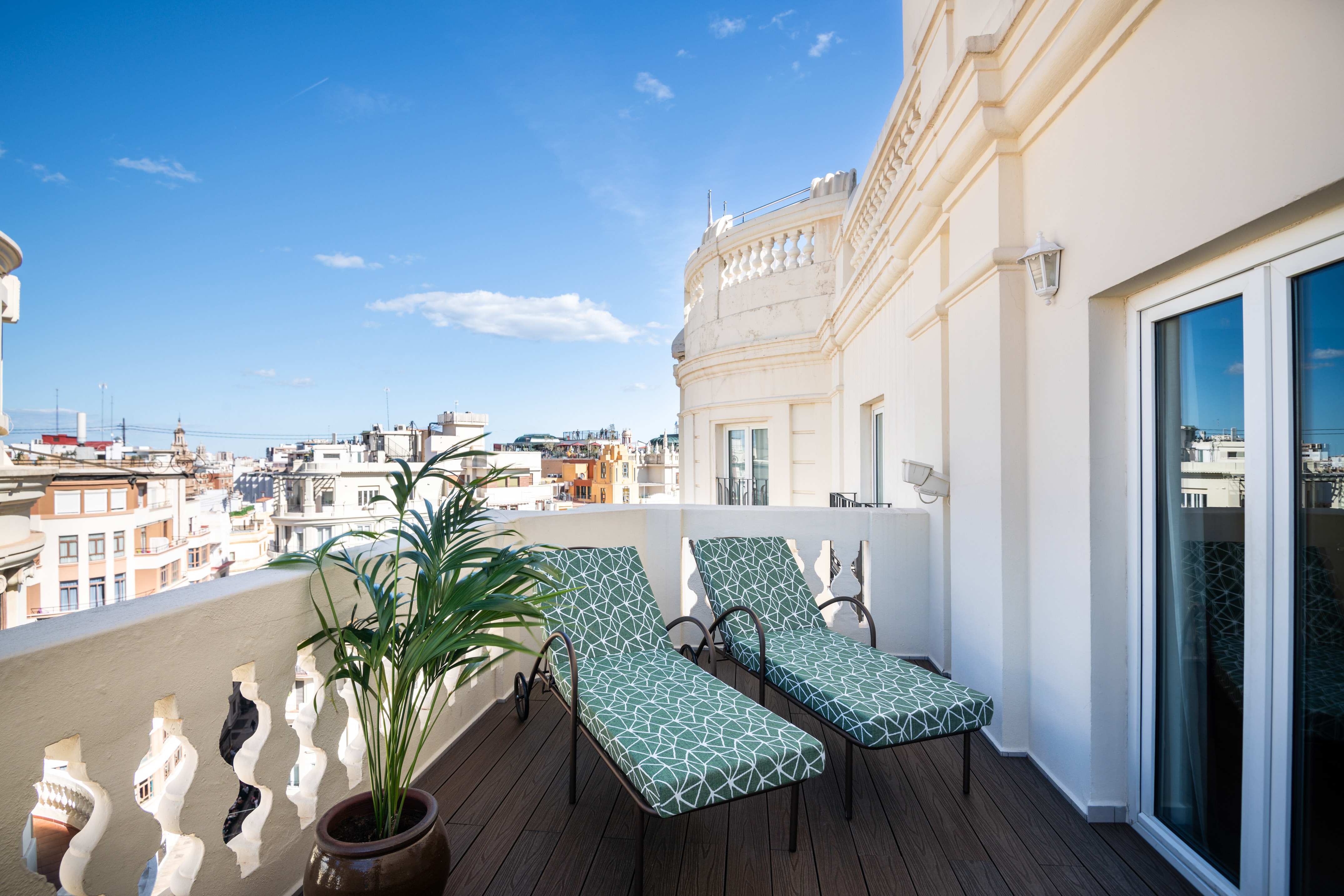 Sunny rooftop terrace with green lounge chairs, potted plant, and city skyline.