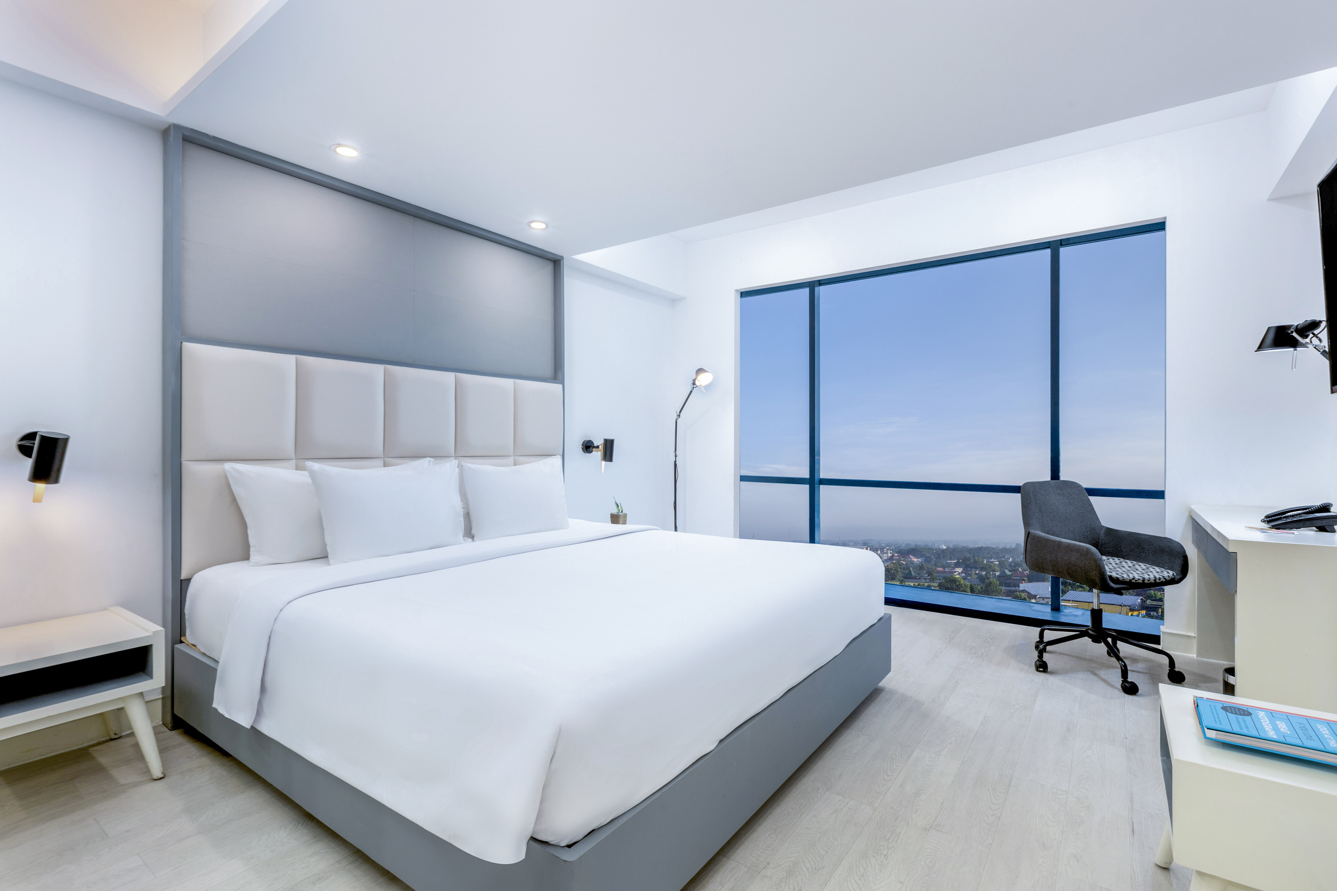 Modern hotel room with white bed, desk, and panoramic city view window.
