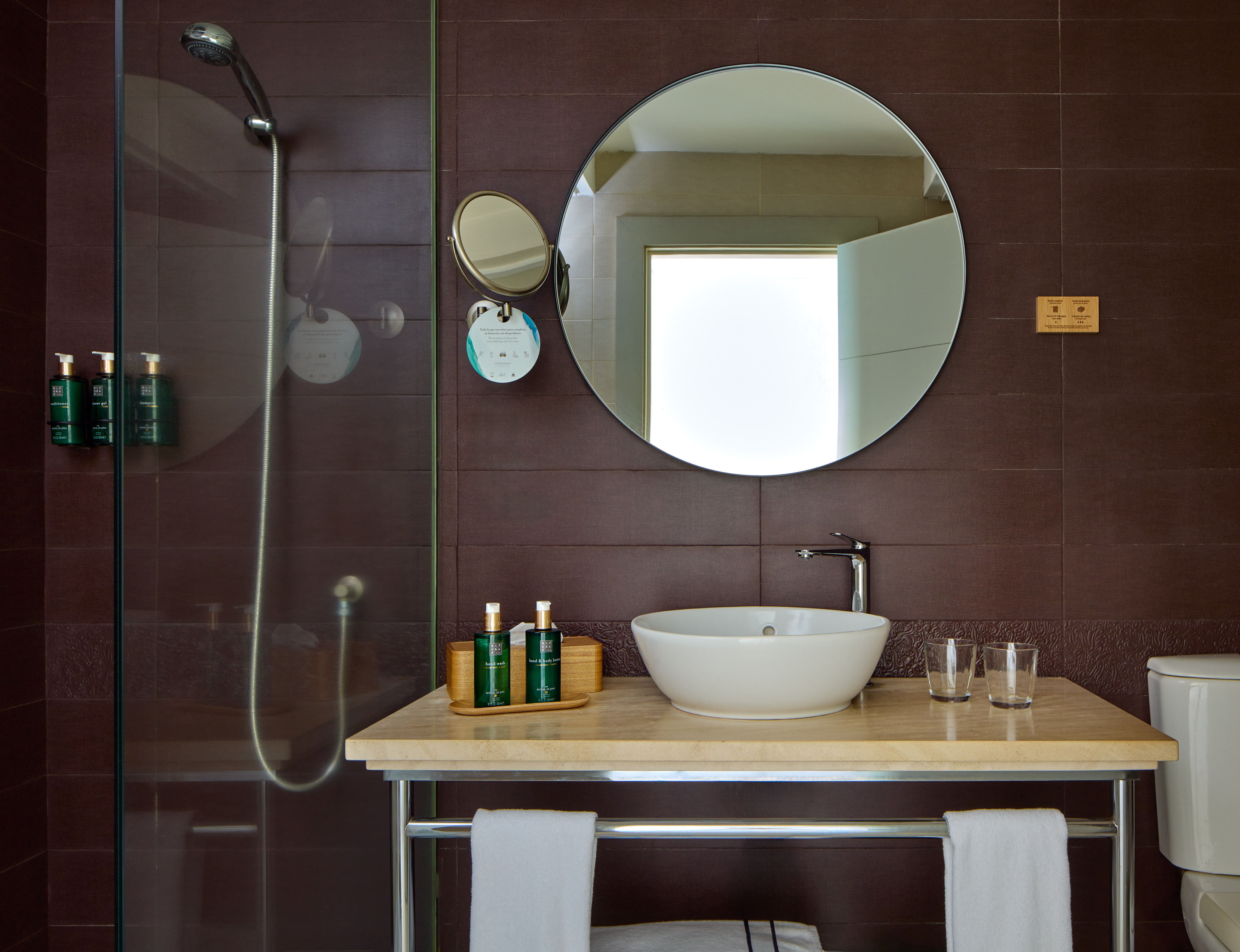 Modern bathroom with brown tiles, glass shower, bowl sink, and round mirror.