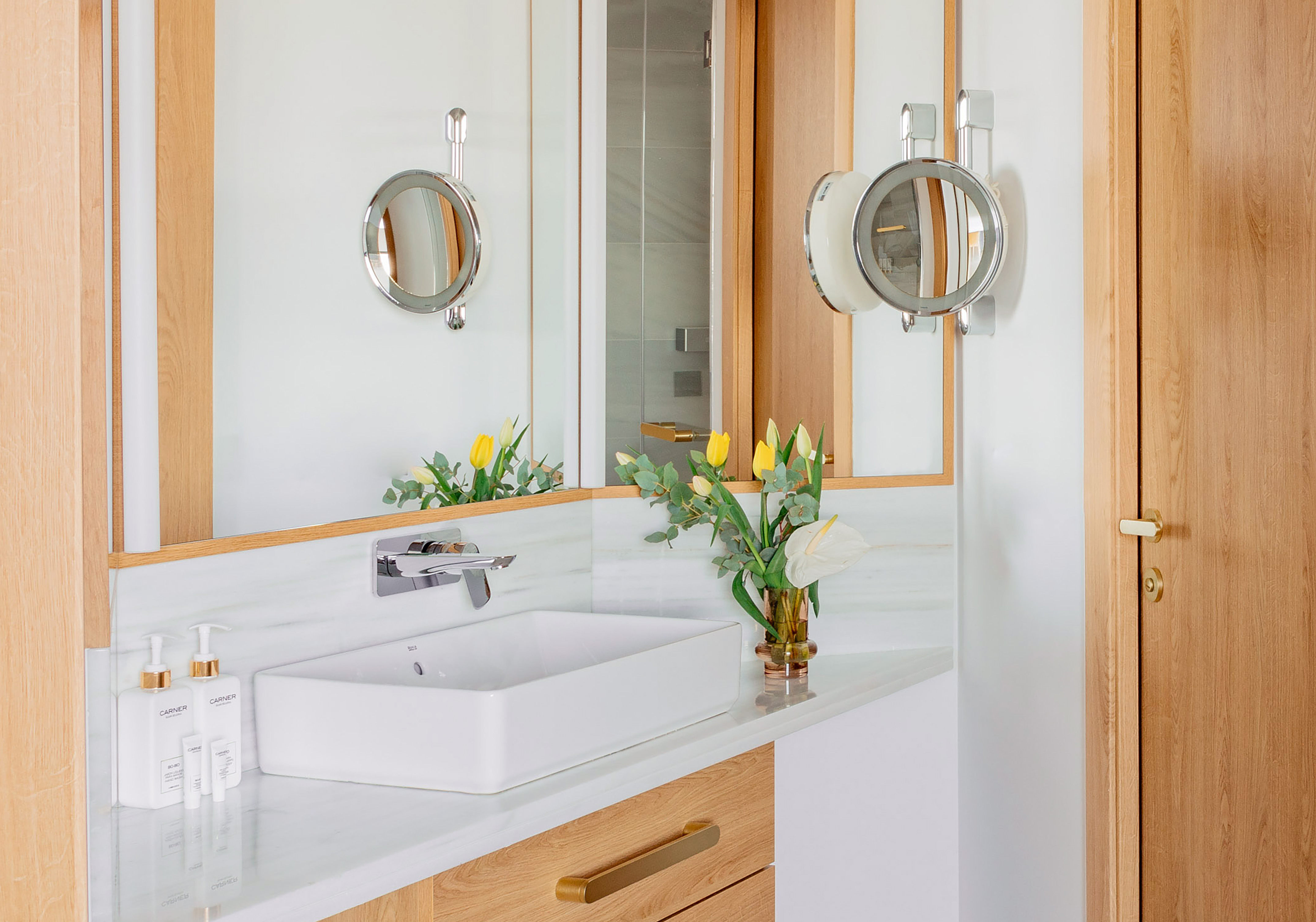 Modern bathroom vanity with white sink, marble counter, wood, lighted mirrors.