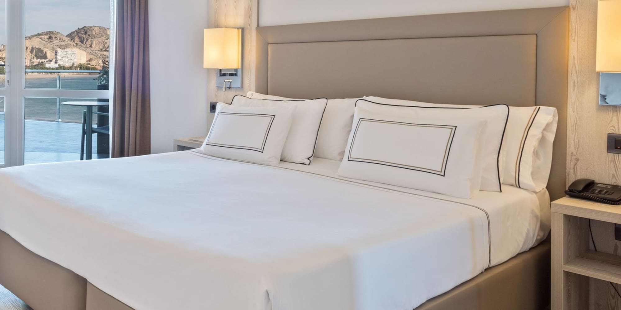 Habitación de hotel moderna con cama doble, vista al mar y montaña desde balcón.
