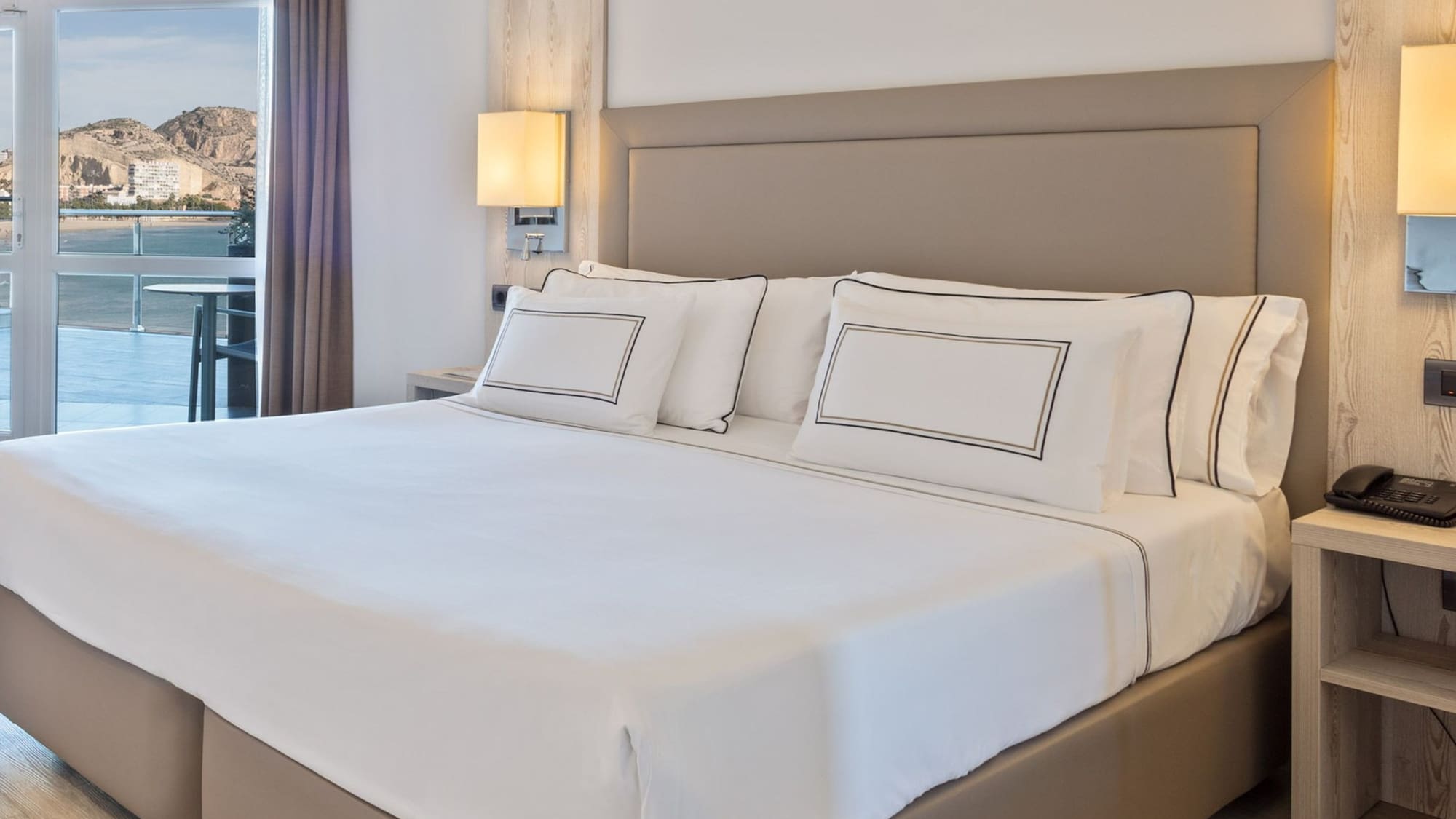 Habitación de hotel moderna con cama doble, vista al mar y montaña desde balcón.