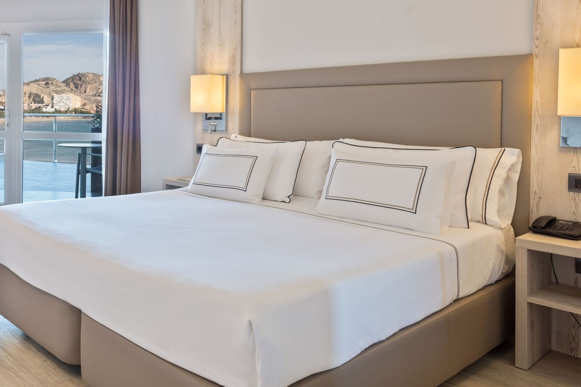 Dormitorio hotel moderno con cama, vistas al mar y montaña desde el balcón.