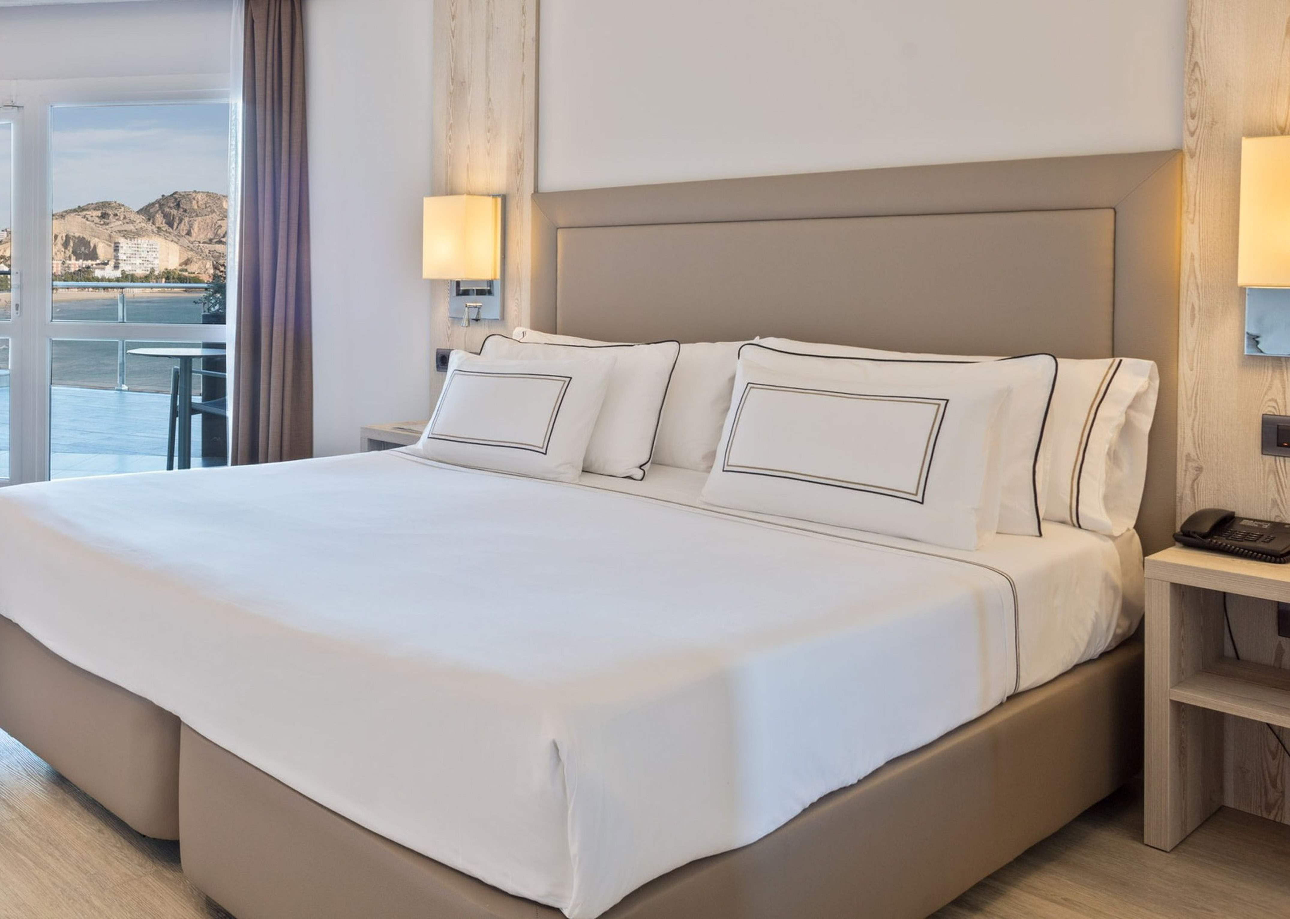 Habitación de hotel moderna con cama doble, vista al mar y montaña desde balcón.