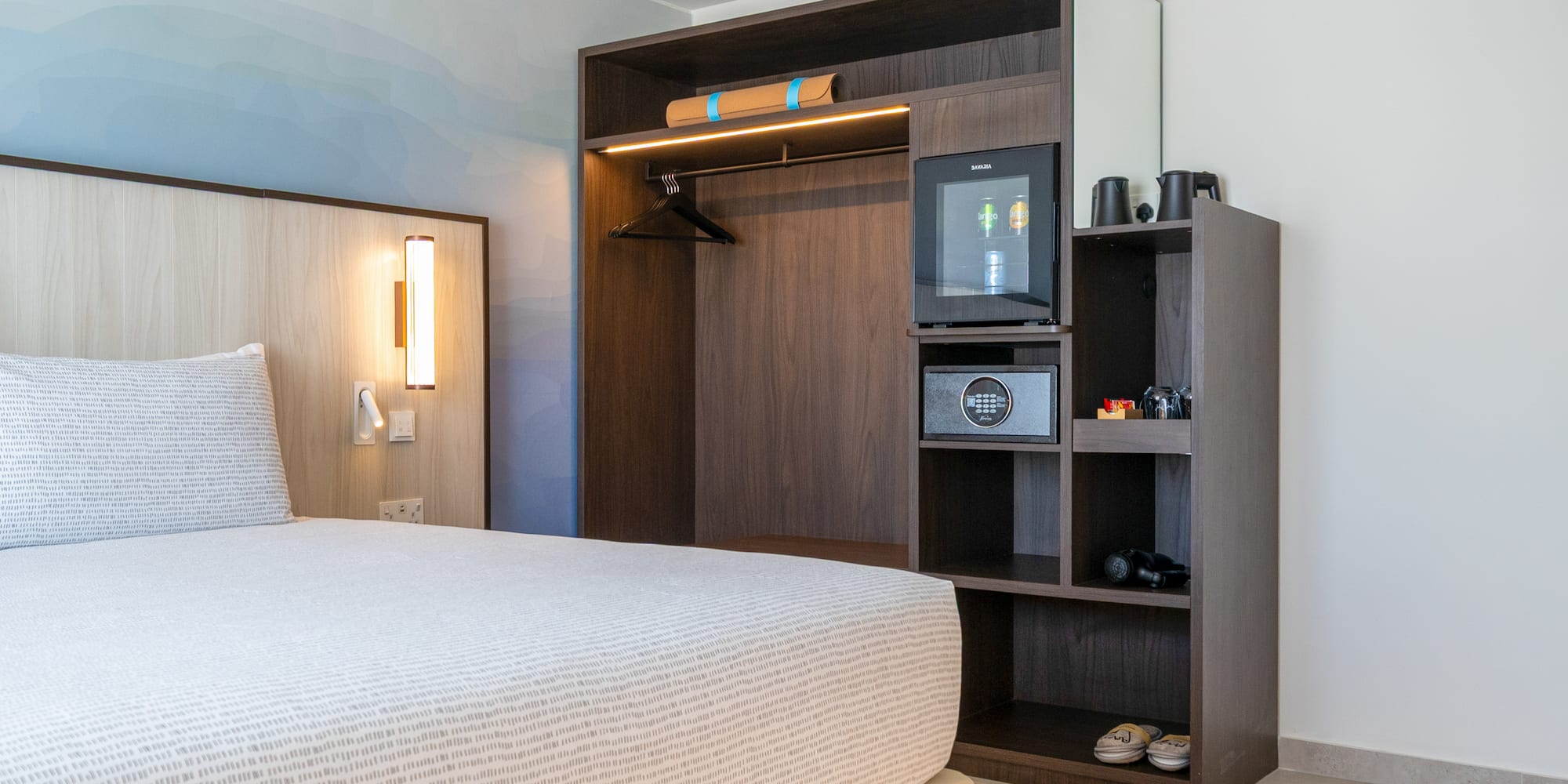 Modern hotel bedroom with built-in wardrobe, minibar, safe, and patterned bed.