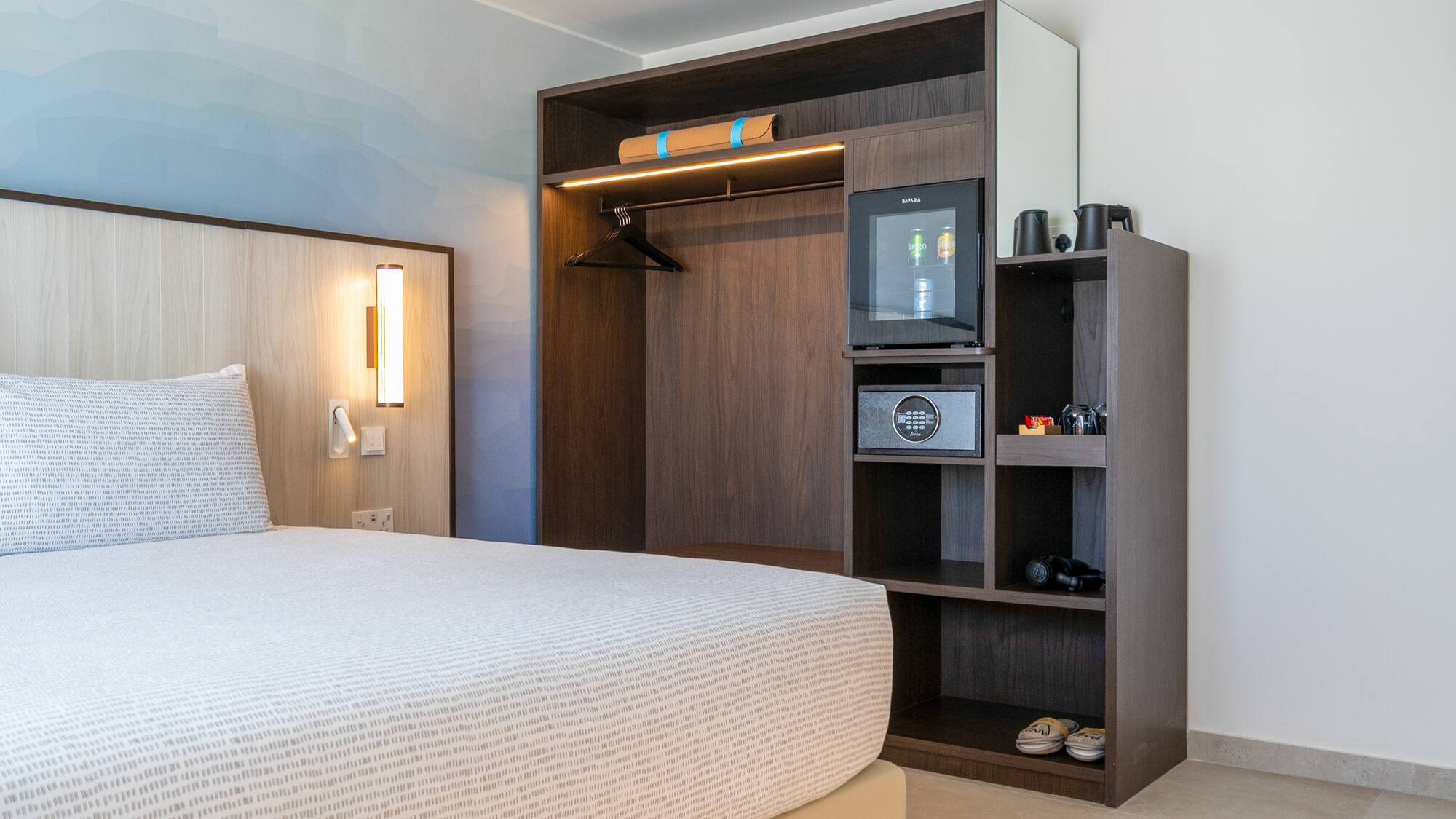 Modern hotel bedroom with built-in wardrobe, minibar, safe, and patterned bed.
