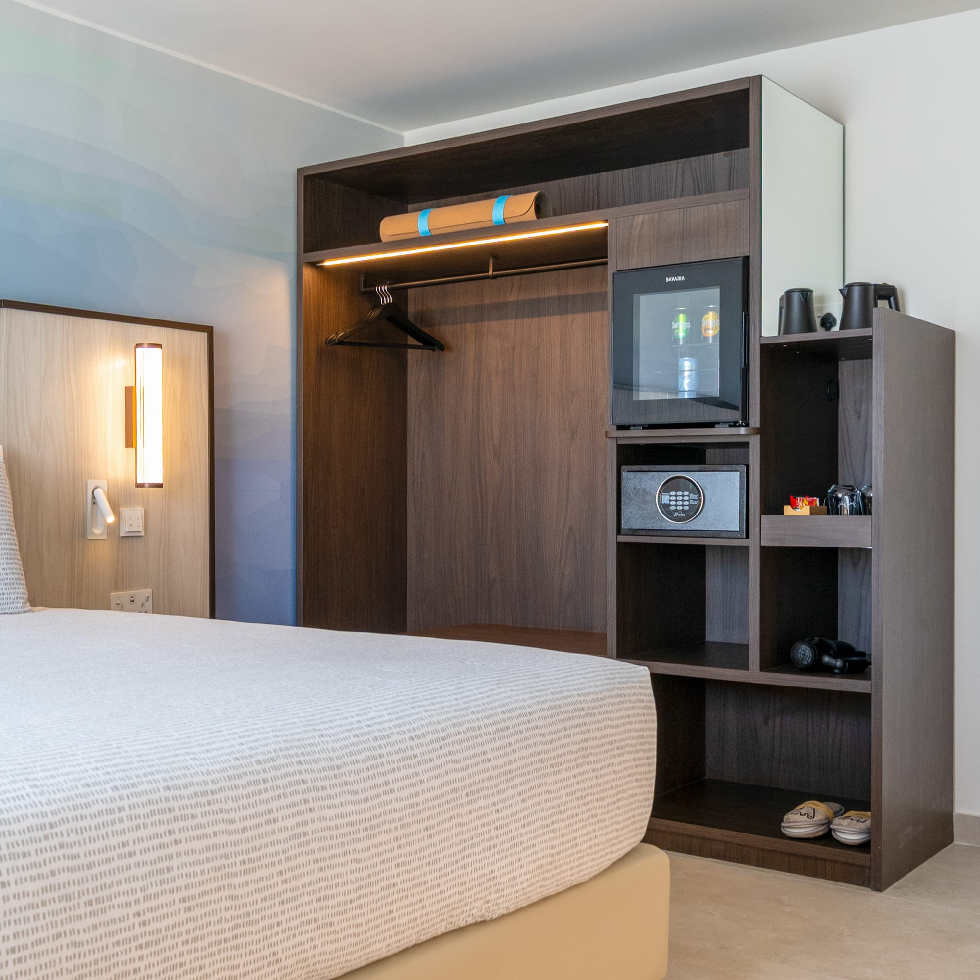 Modern hotel bedroom with built-in wardrobe, minibar, safe, and patterned bed.