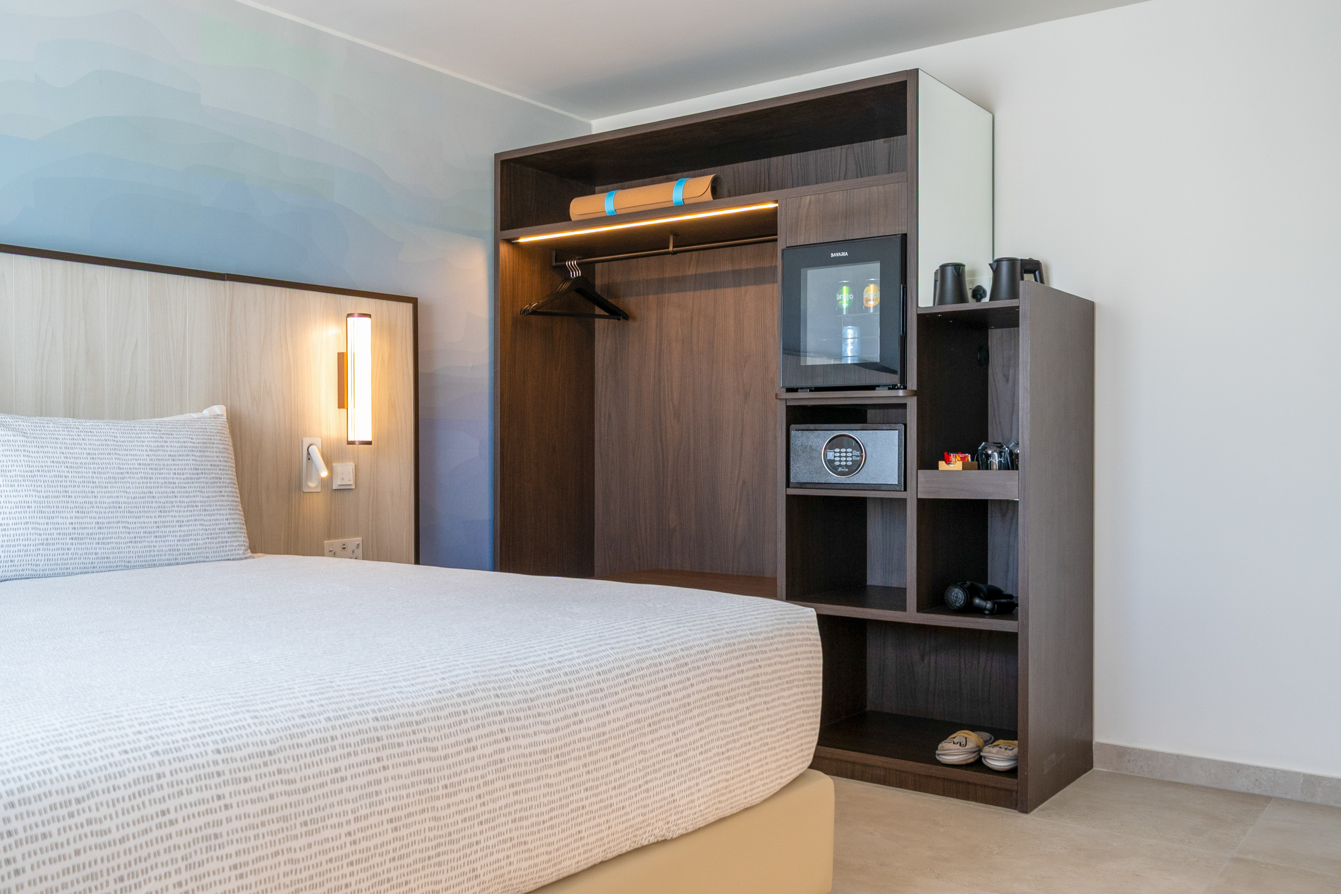 Modern hotel bedroom with built-in wardrobe, minibar, safe, and patterned bed.