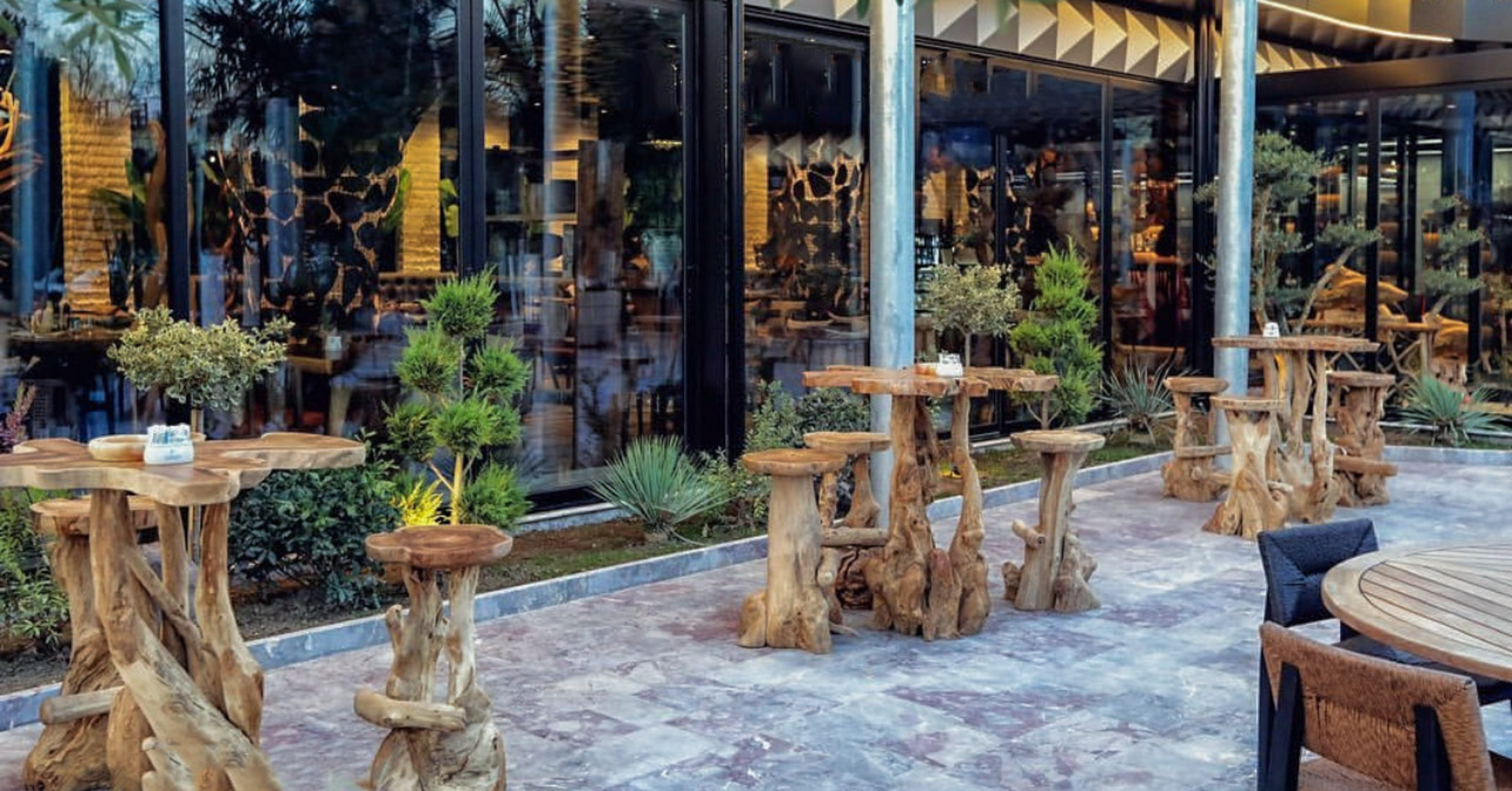 Chic outdoor dining patio with natural wood tables, stools, and greenery.