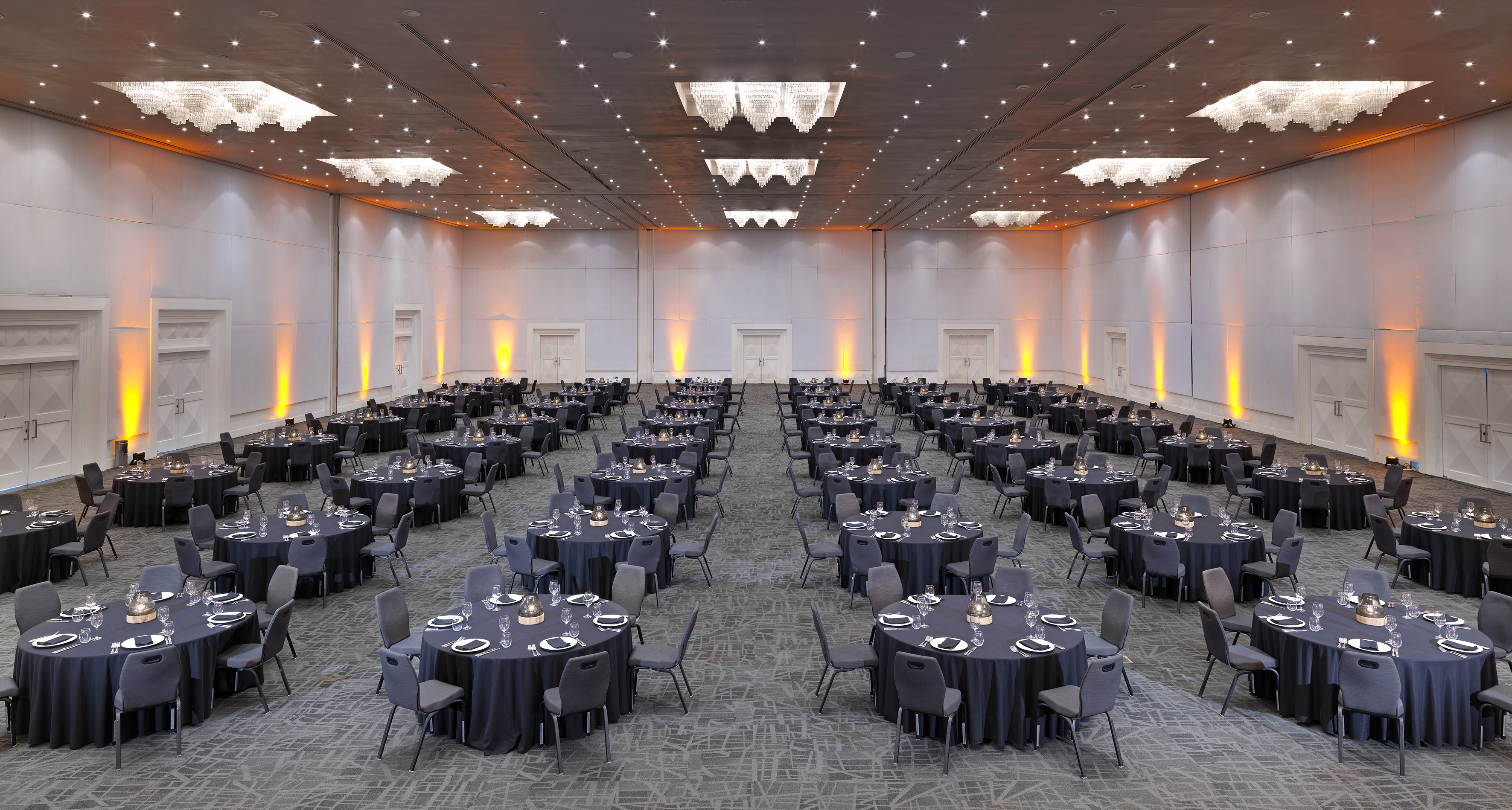 Grand ballroom with dark banquet setup, chandeliers, and orange wall uplighting.