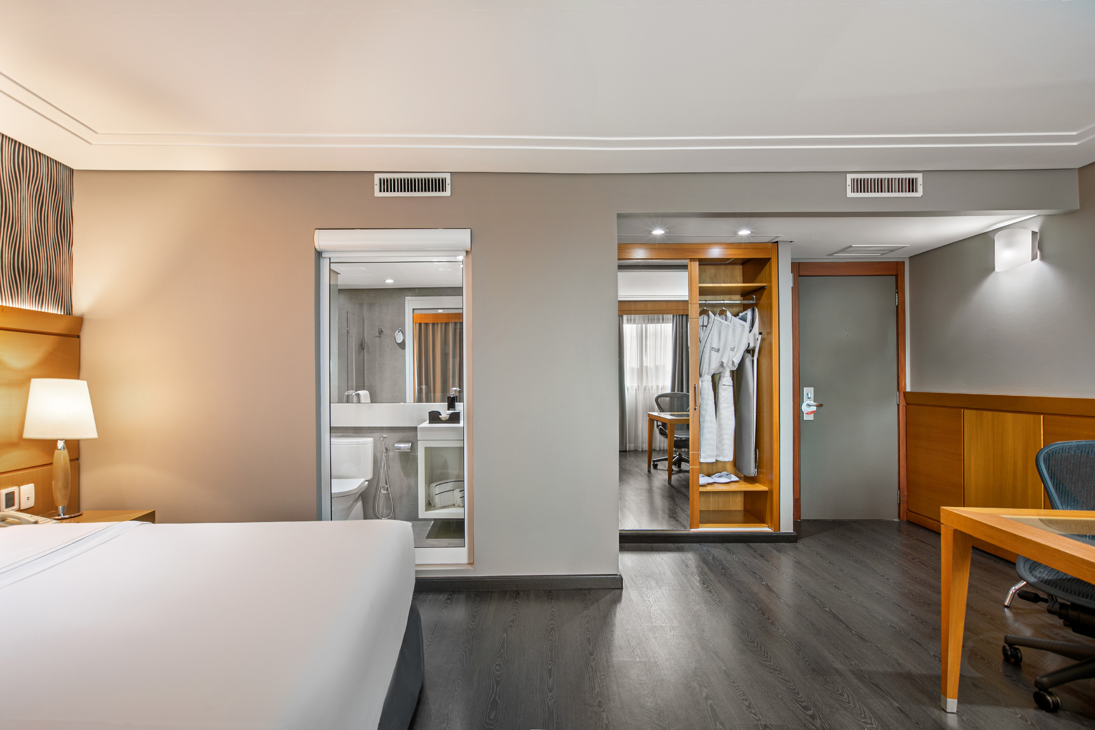 Modern hotel room interior with bed, open bathroom, wardrobe, and work desk.