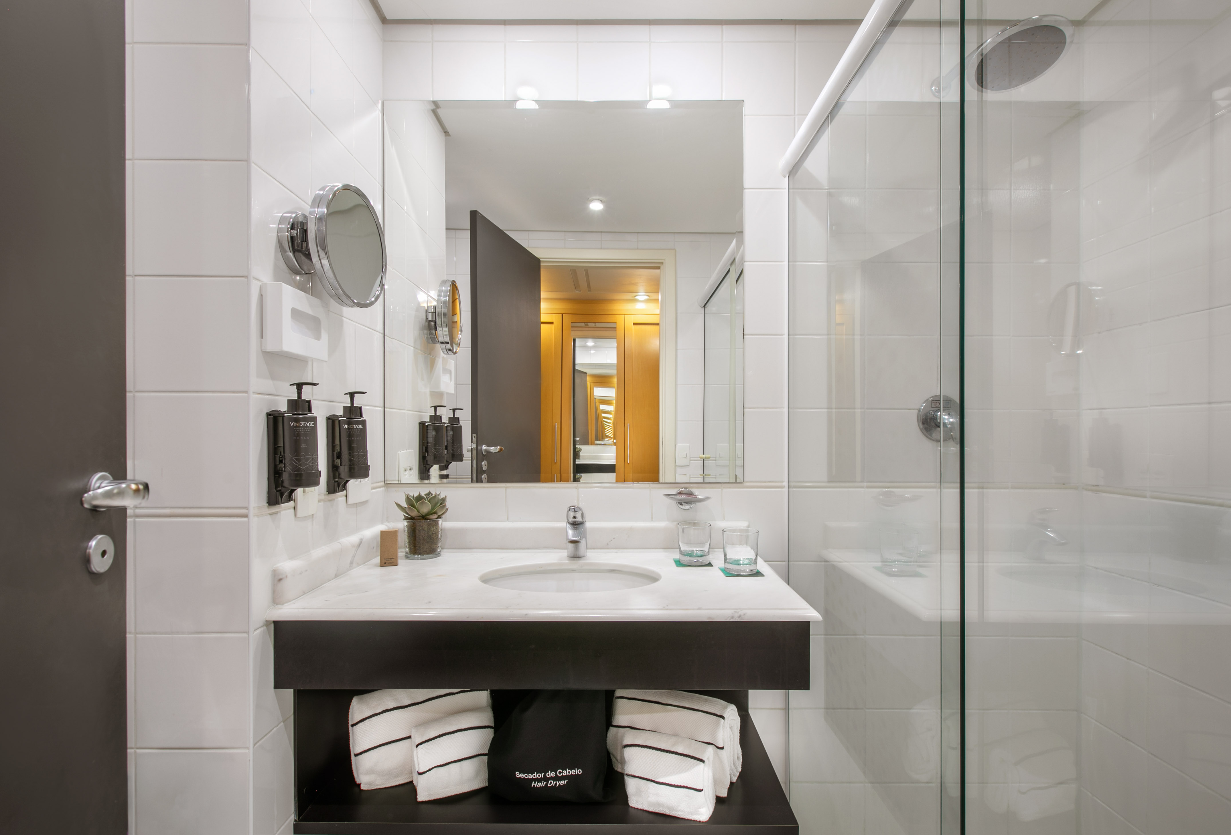White tiled hotel bathroom with marble vanity, sink, mirror, and glass shower.