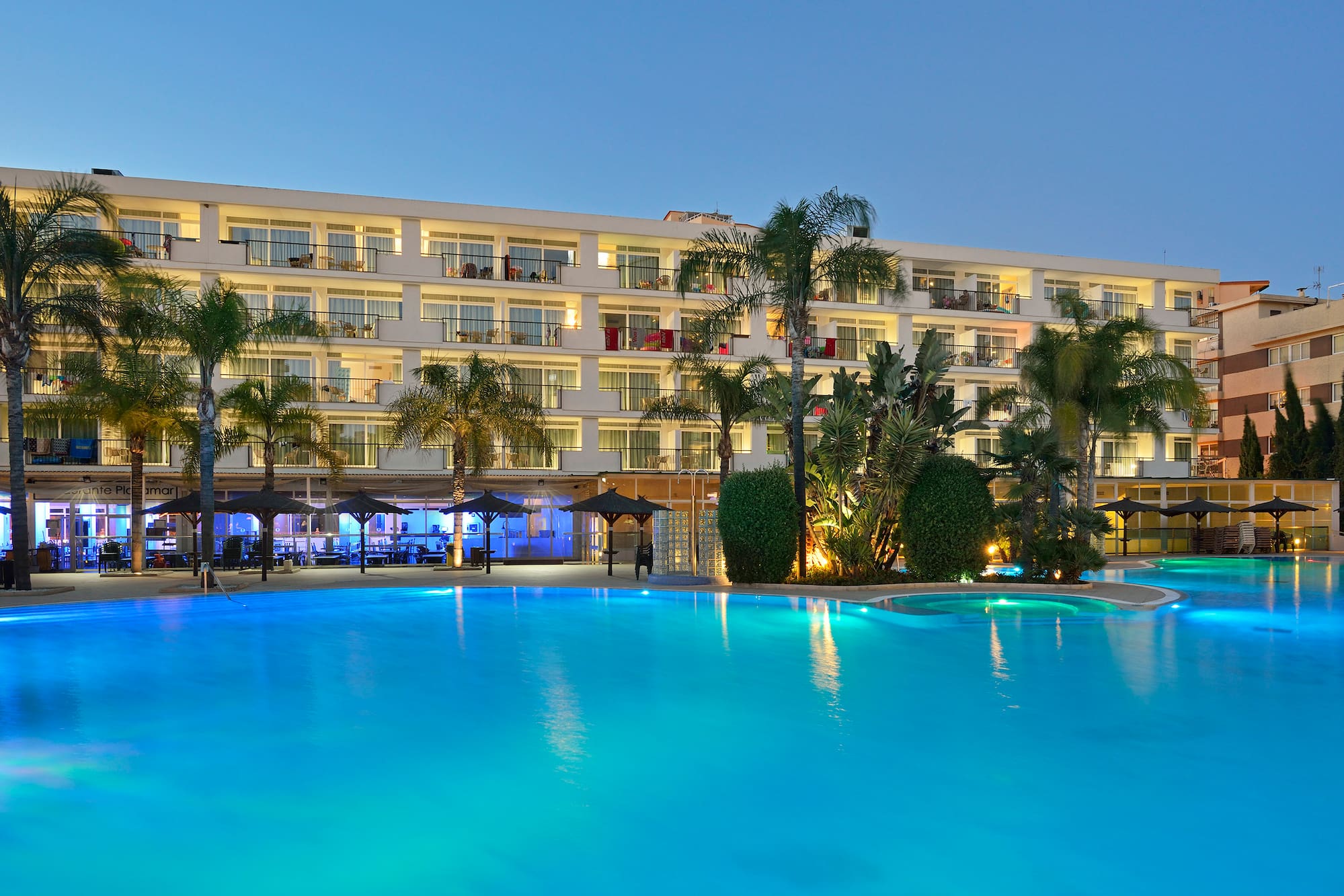 Hotel con piscina iluminada de azul al anochecer, palmeras y balcones.