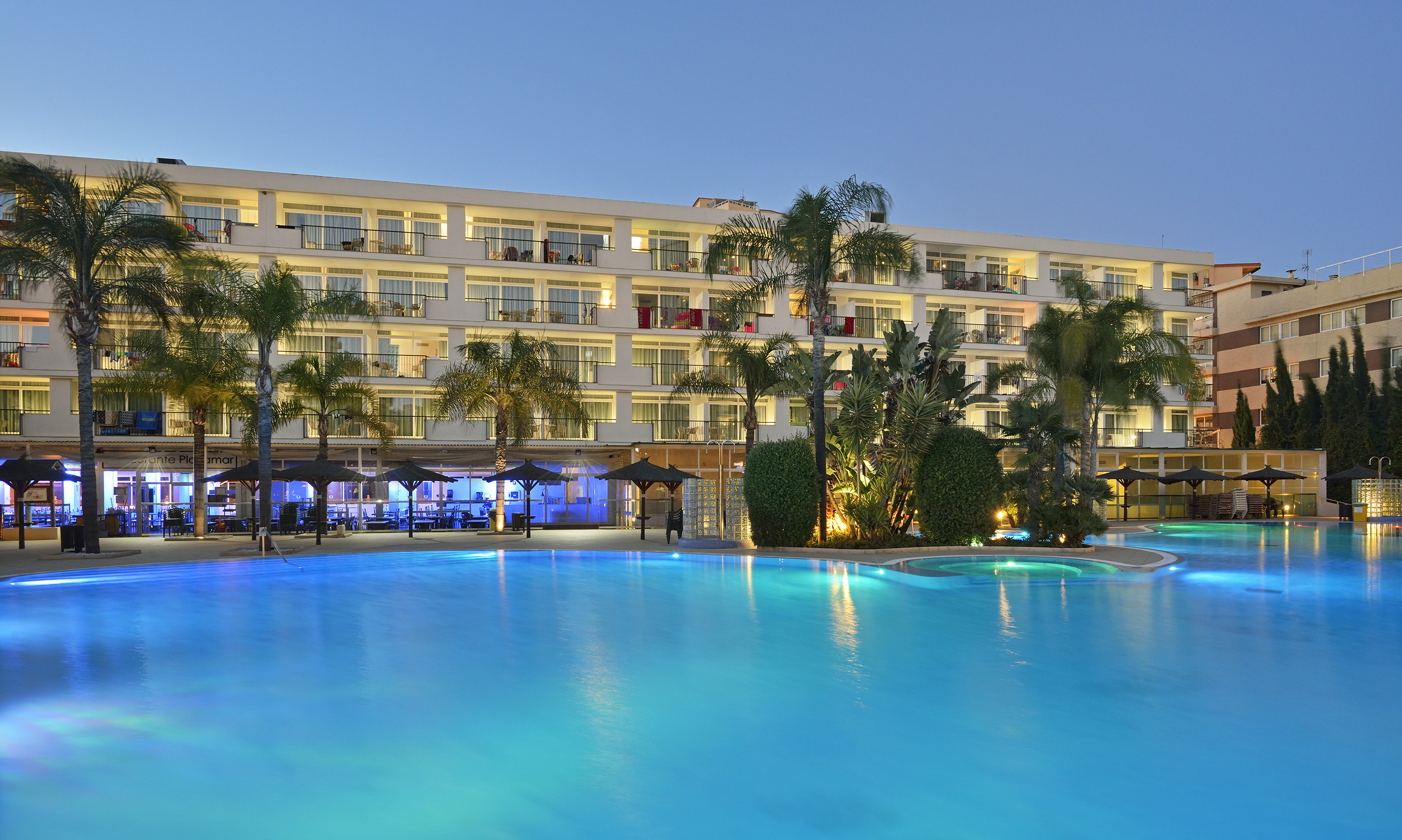 Hotel con piscina iluminada de azul al anochecer, palmeras y balcones.