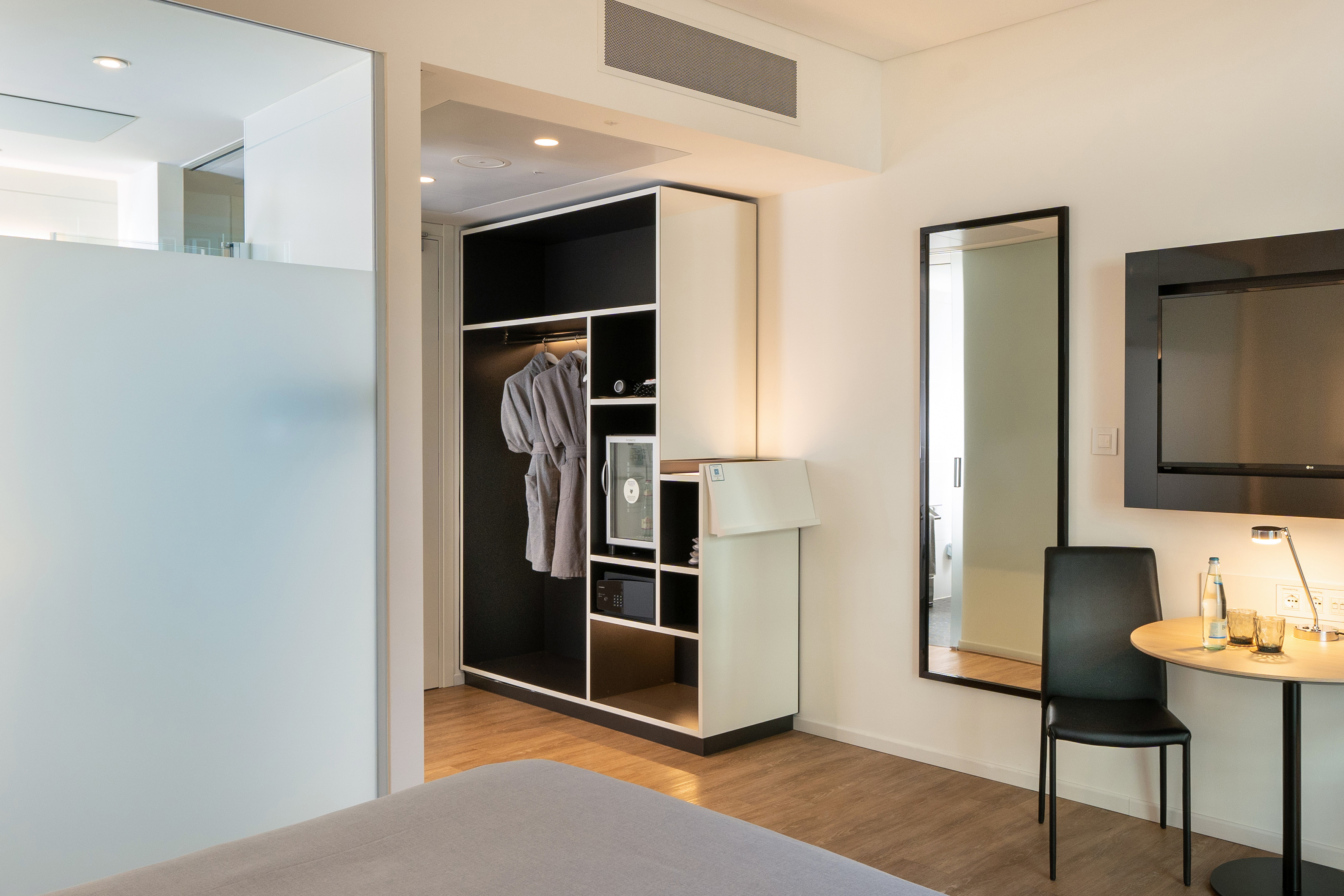 Modern hotel room with open wardrobe, bathrobes, minibar, safe, and mirror.