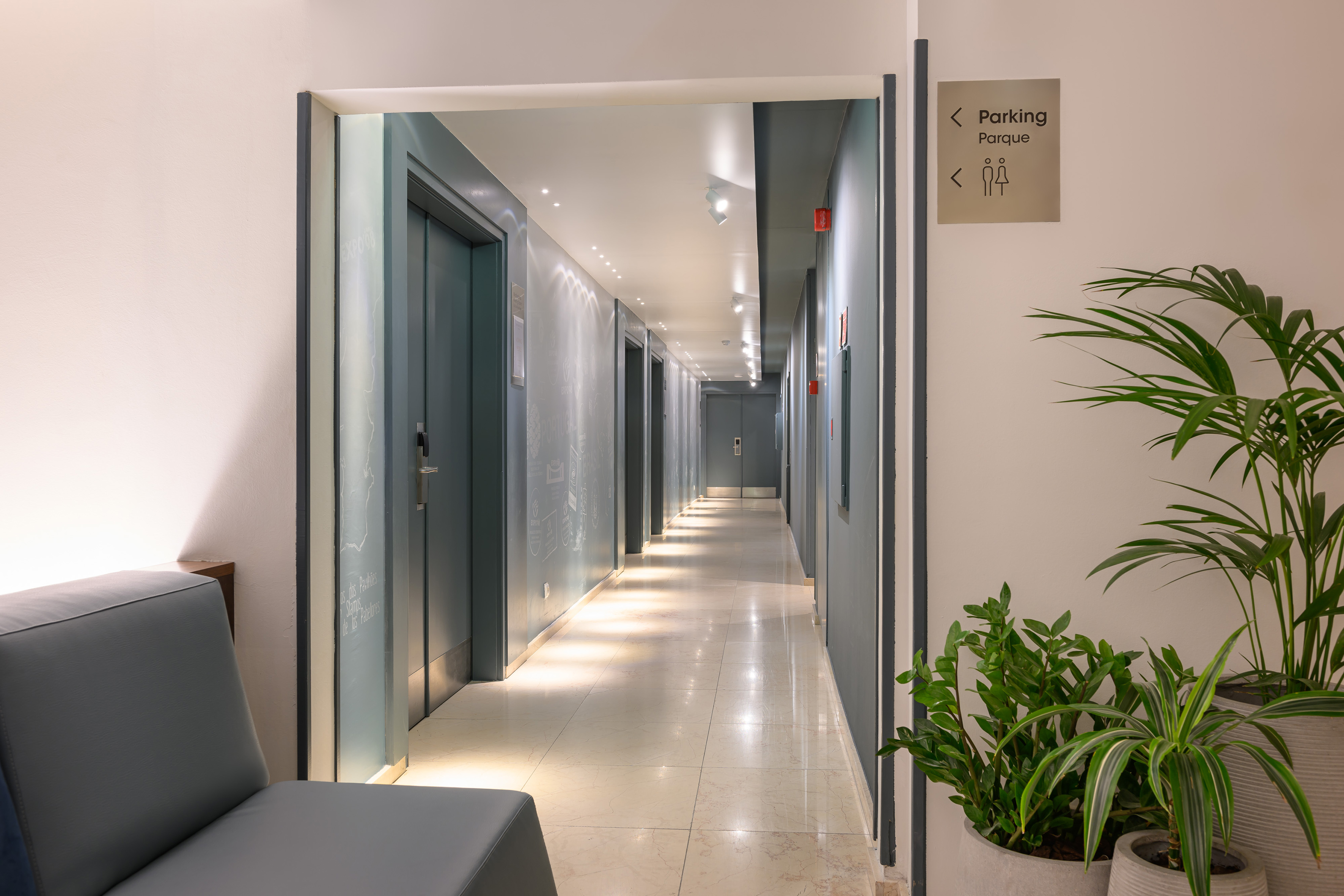 Bright hotel corridor with elevator access, lounge area, and potted plants.