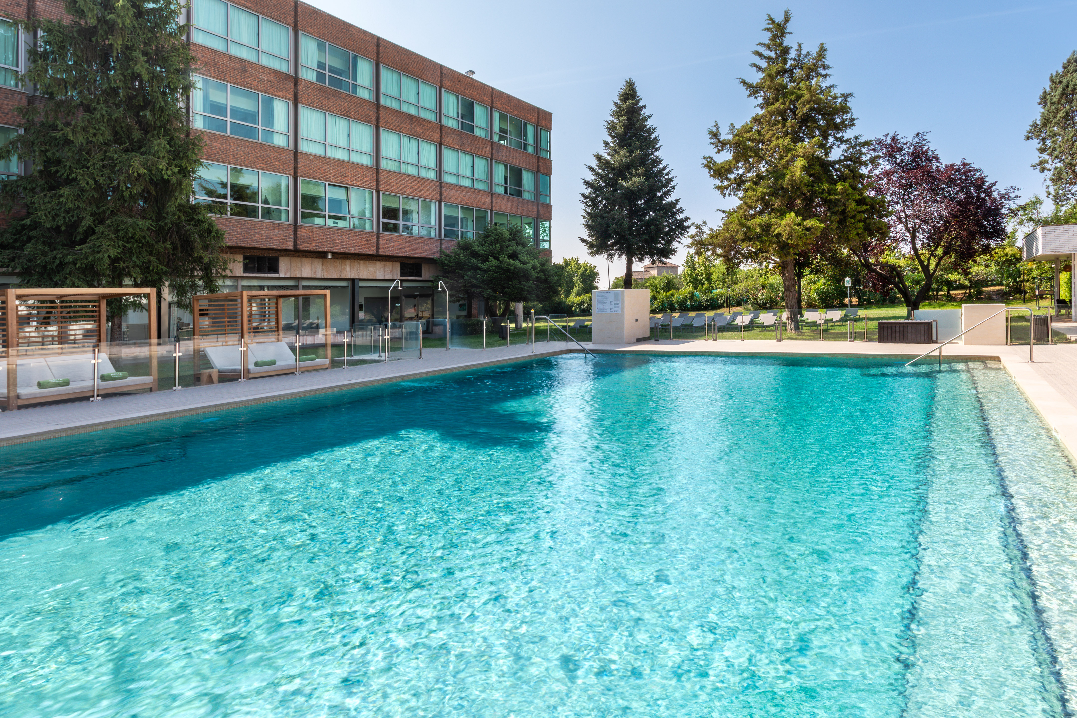 Outdoor hotel swimming pool, sun loungers, brick building, and garden.