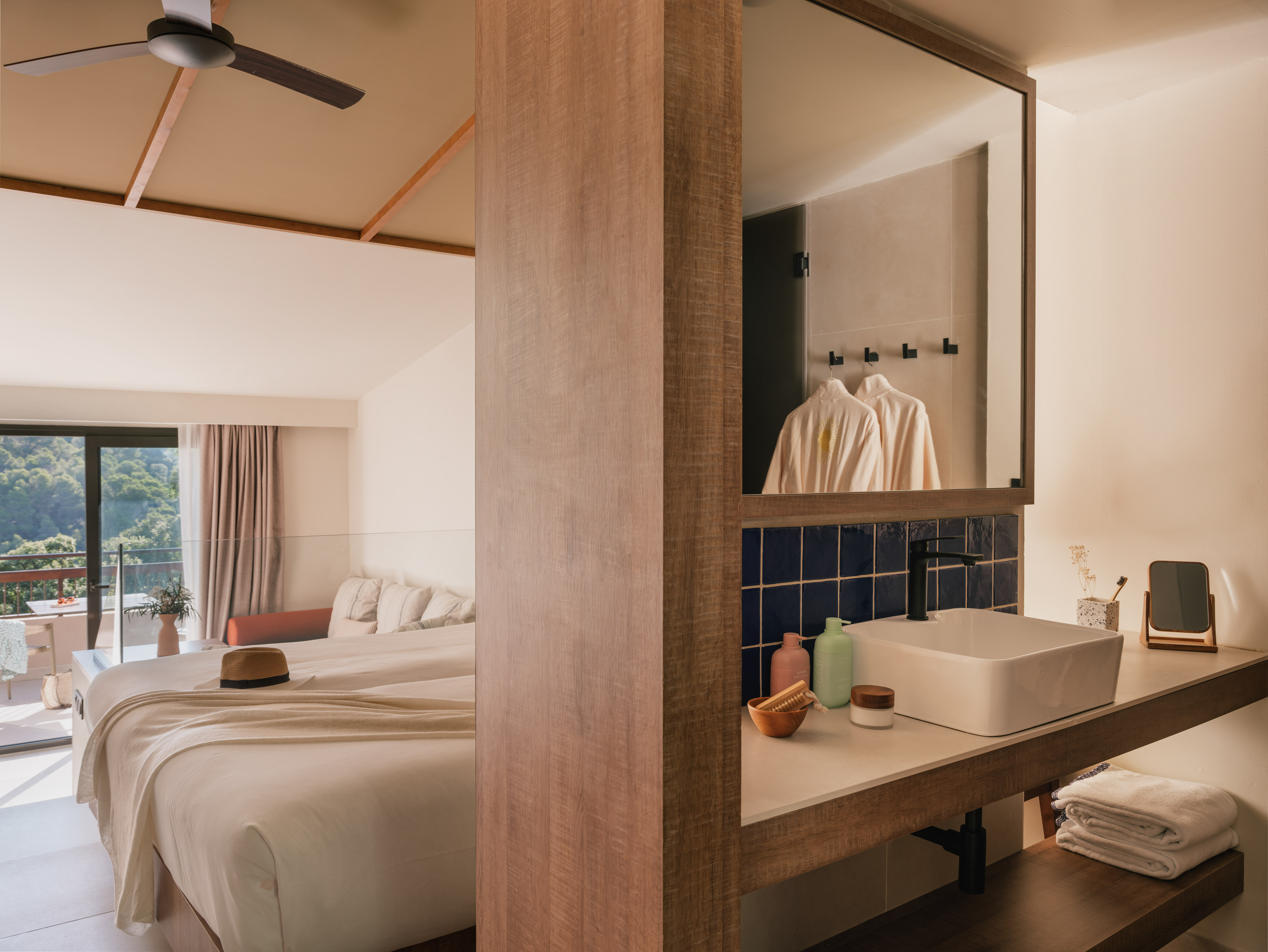 Modern hotel room with bed, balcony nature view, and open-plan bathroom with sink.