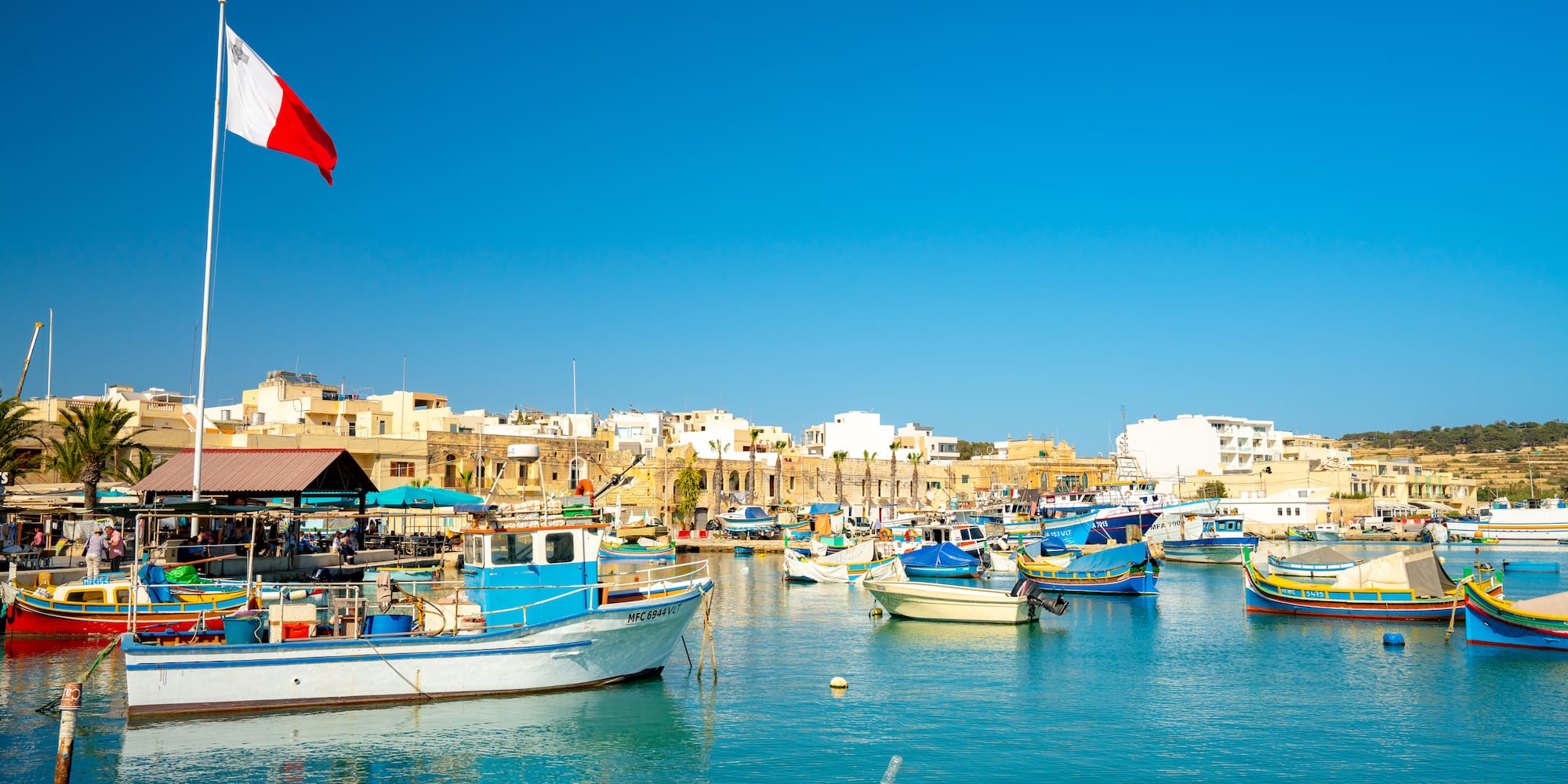 Maltese flag, colorful fishing boats in Marsaxlokk harbor under clear blue sky.