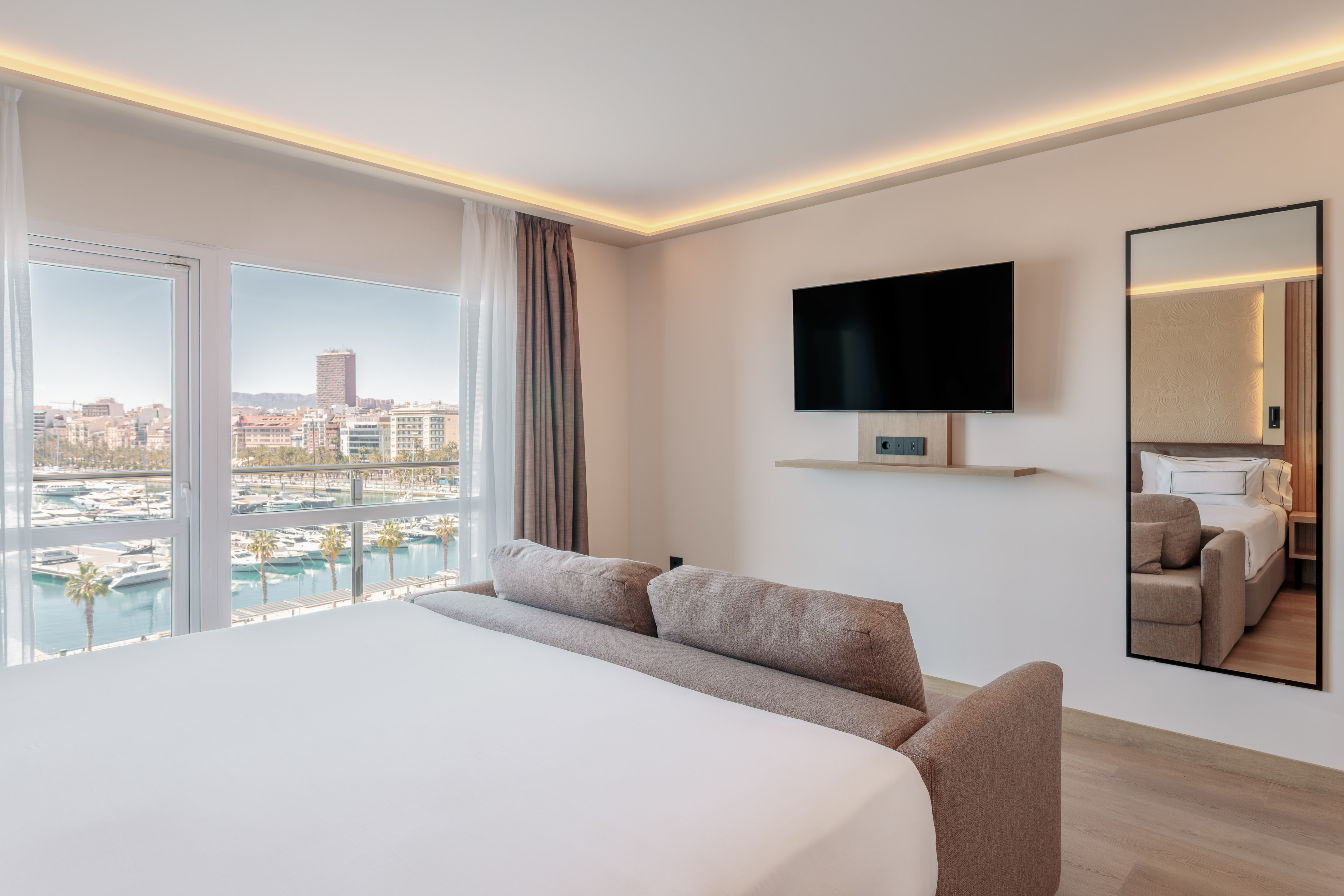 Bright hotel suite with sofa bed, wall TV, and marina port view.