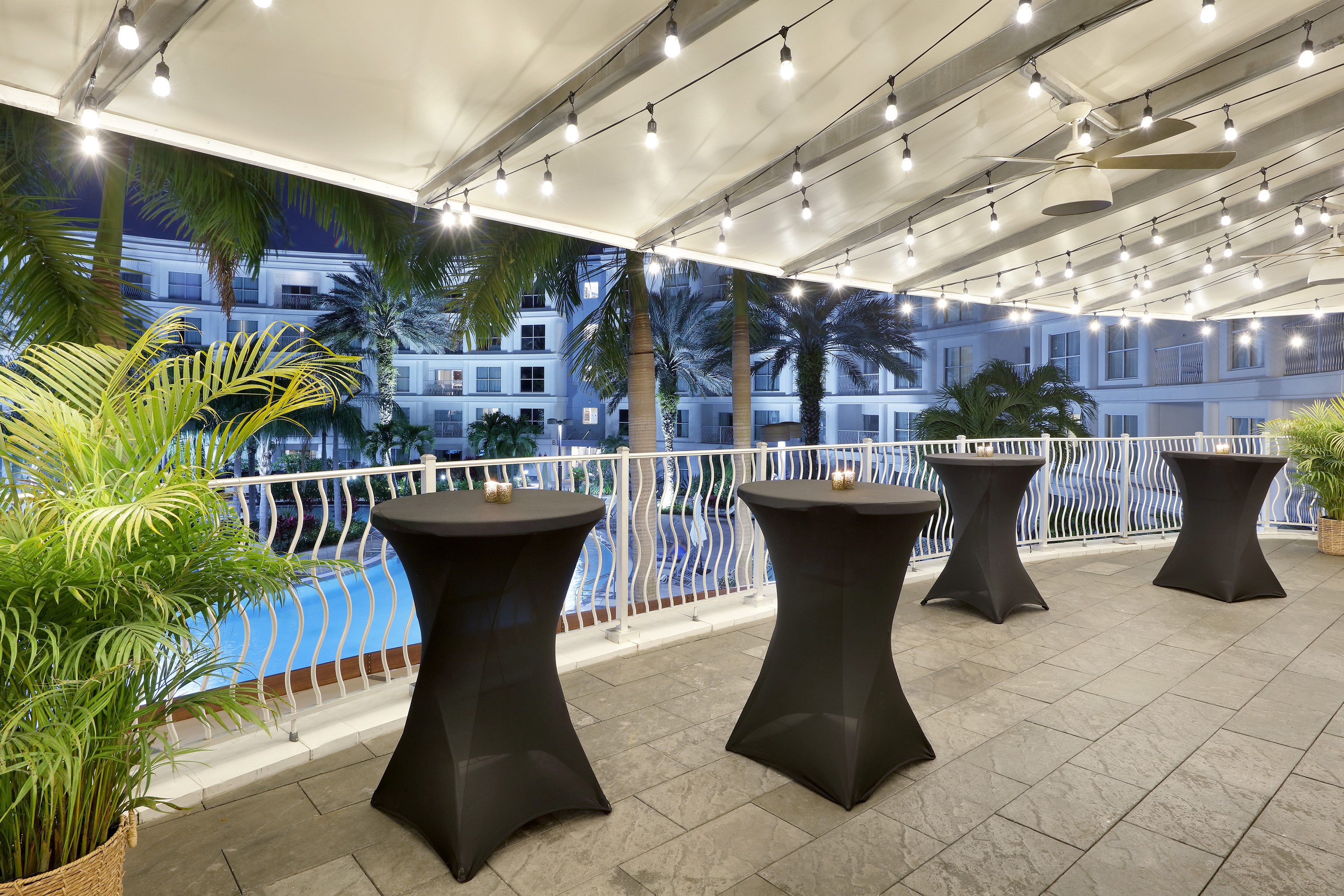 Evening resort patio with string lights, cocktail tables, palm trees, and pool.