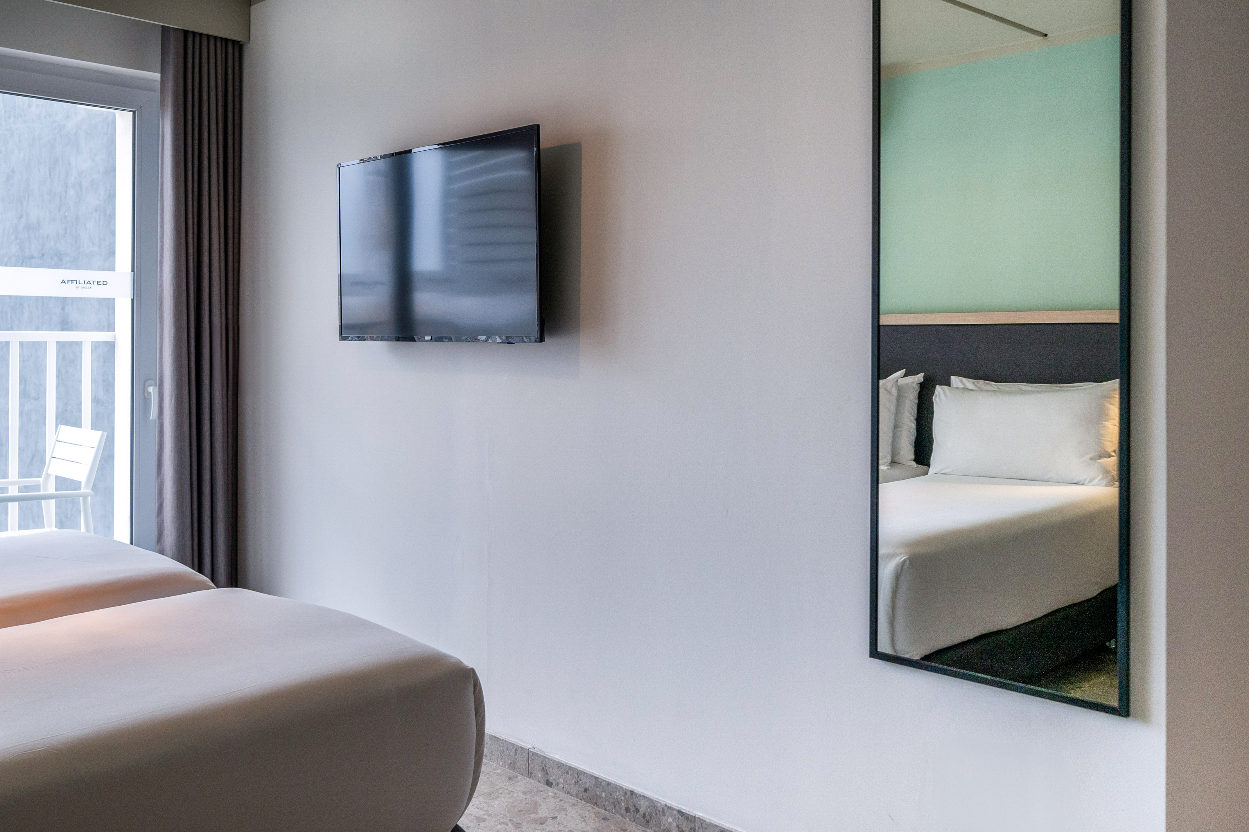 Modern hotel room with twin beds, wall TV, mirror, and balcony.