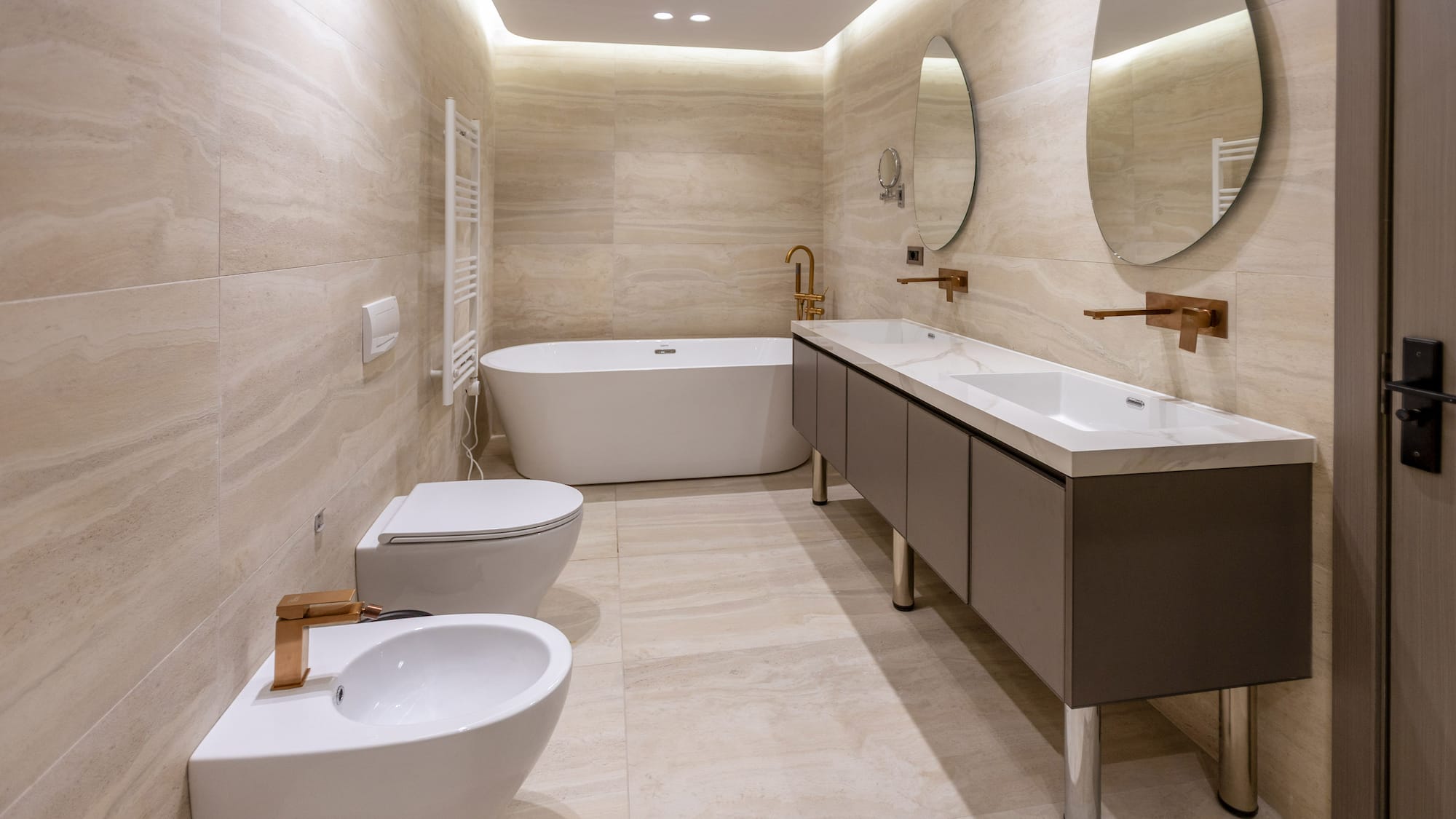 Modern beige marble bathroom with freestanding tub, double vanity, copper fixtures.