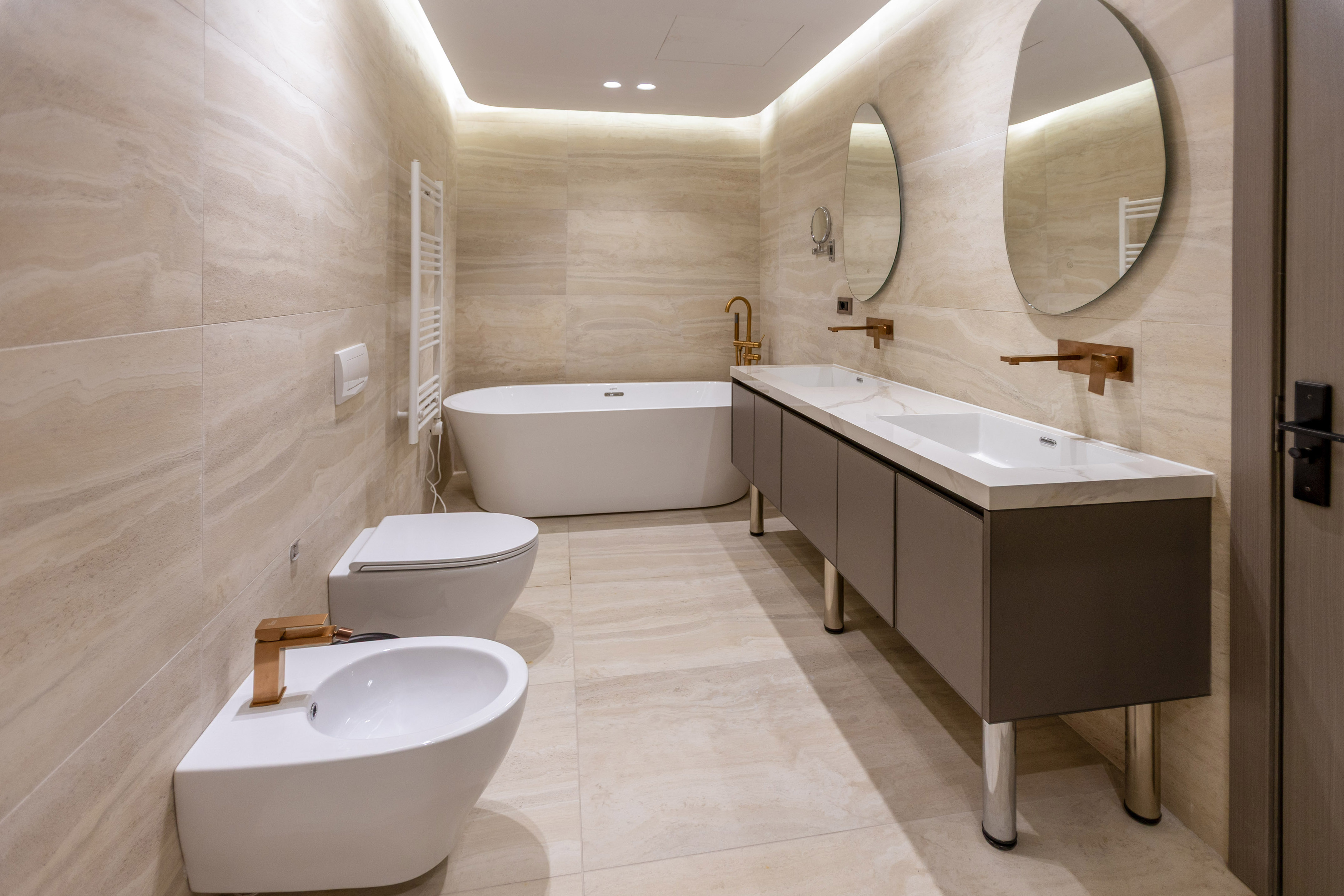 Modern beige marble bathroom with freestanding tub, double vanity, copper fixtures.