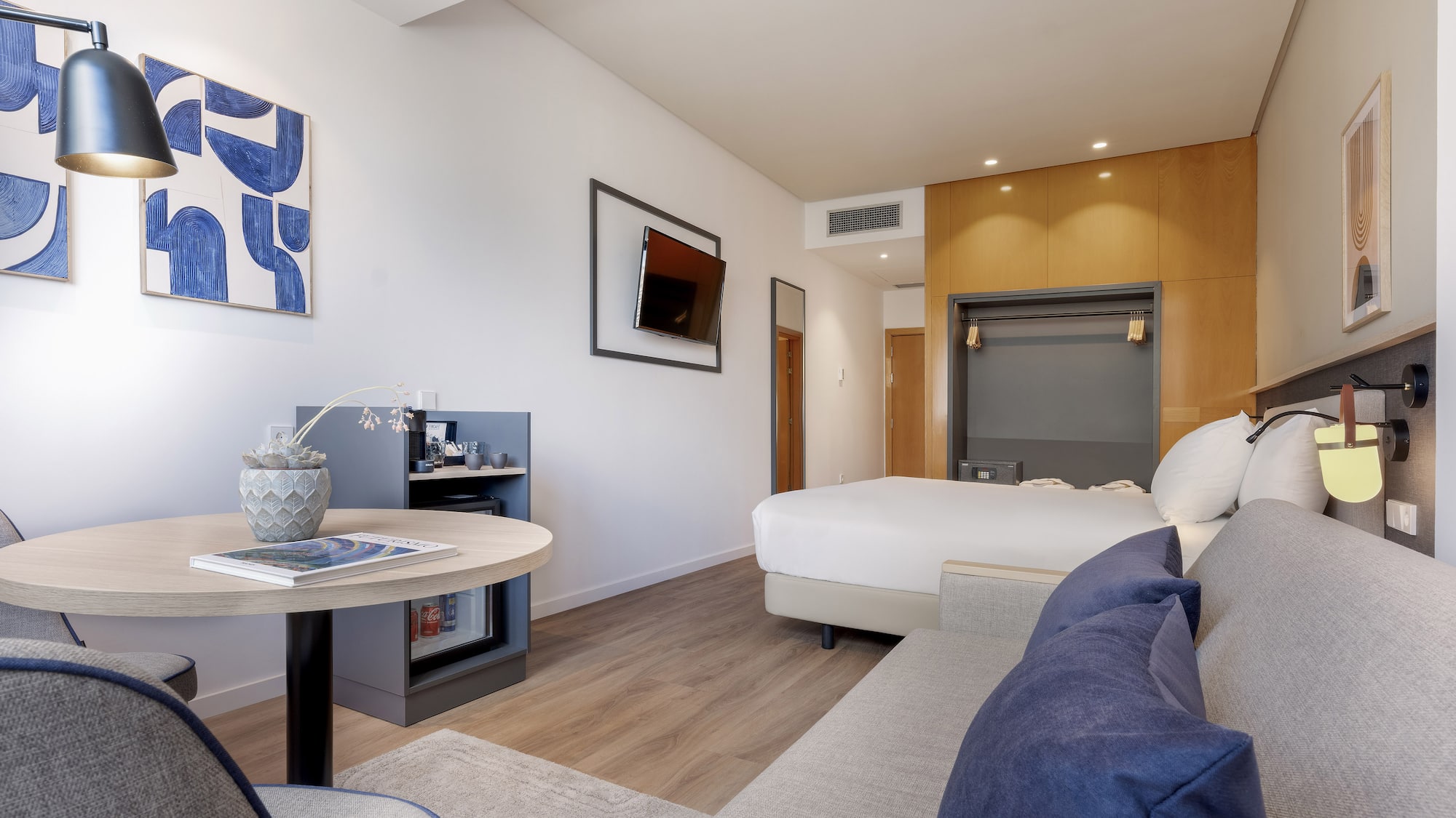 Modern hotel room with bed, grey sofa, round table, TV, and blue art.